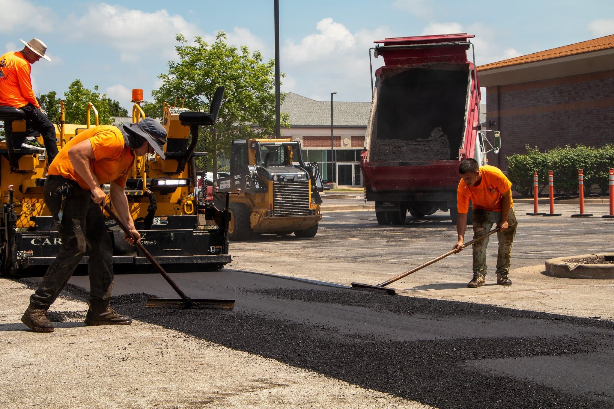 Four workers pave asphalt in front of a Holiday Inn Express hotel on a sunny day, using construction equipment and tools.