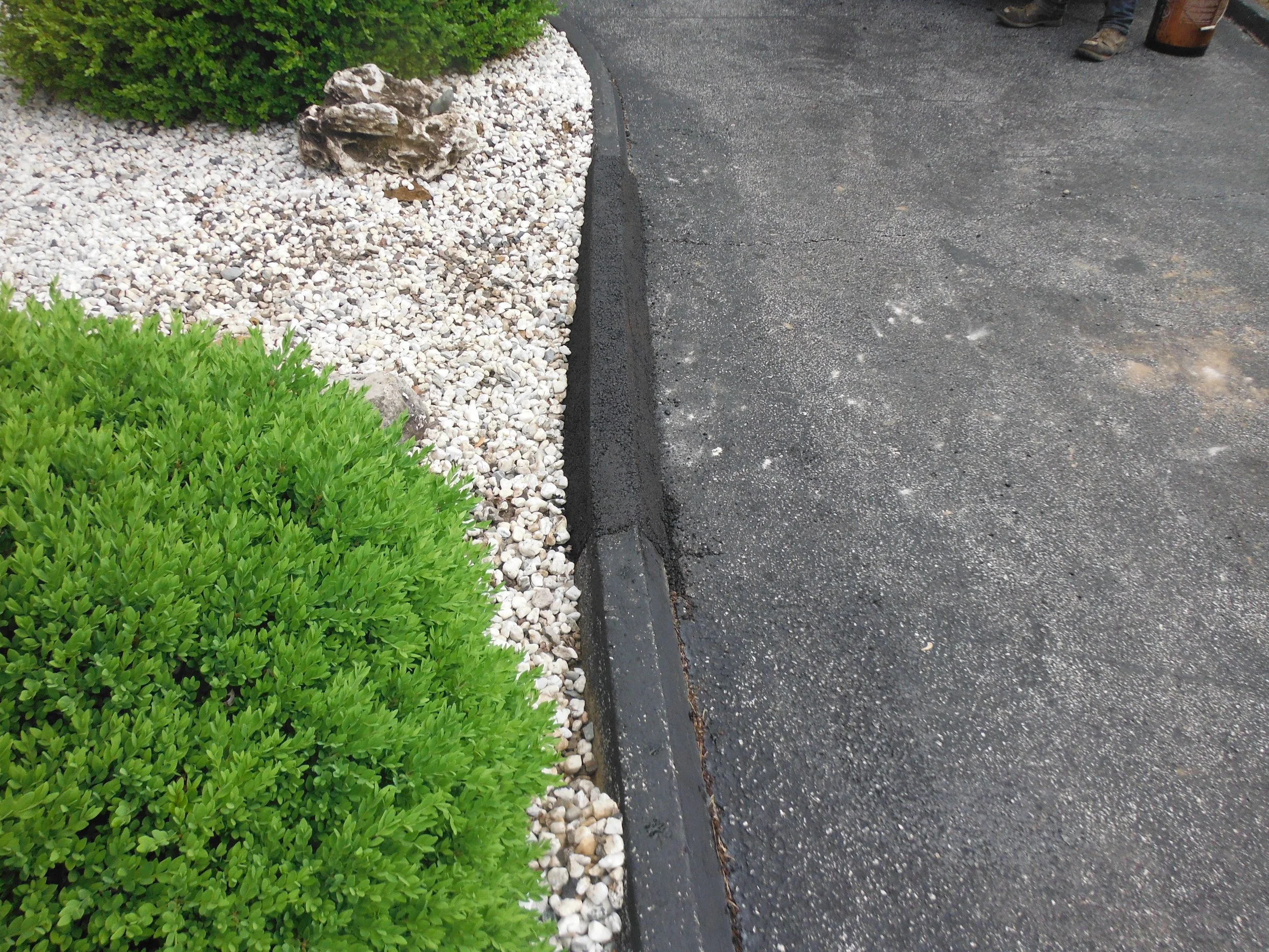 A landscaped area with green bushes, white rocks, and a stone decoration next to a curved curb separating it from a paved asphalt roadway.