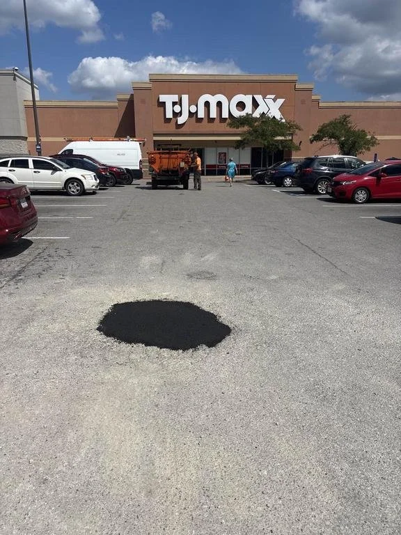 Freshly patched pothole in a parking lot in front of a TJ Maxx store, with cars parked nearby under a partly cloudy sky.