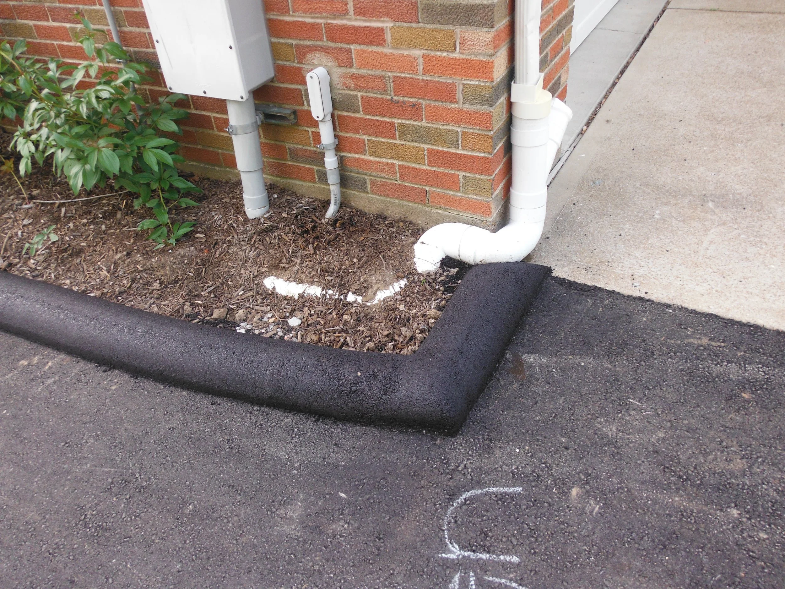 Installation of foam insulation around a pipe at the base of a brick building with an adjacent concrete sidewalk.