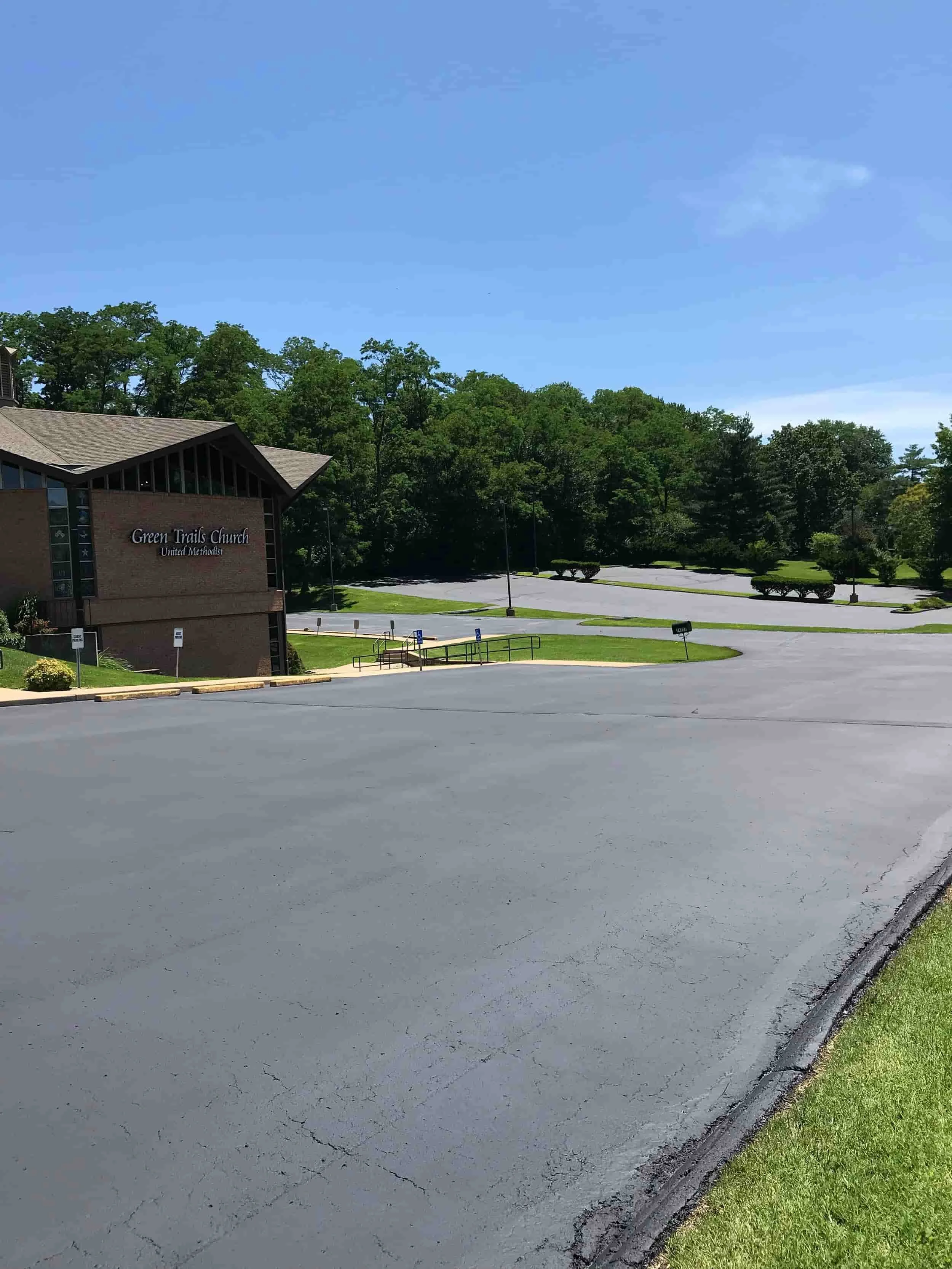 Empty parking lot in front of Green Trails Church, United Methodist, with a building on the left and green trees and bushes in the background, under a clear blue sky.