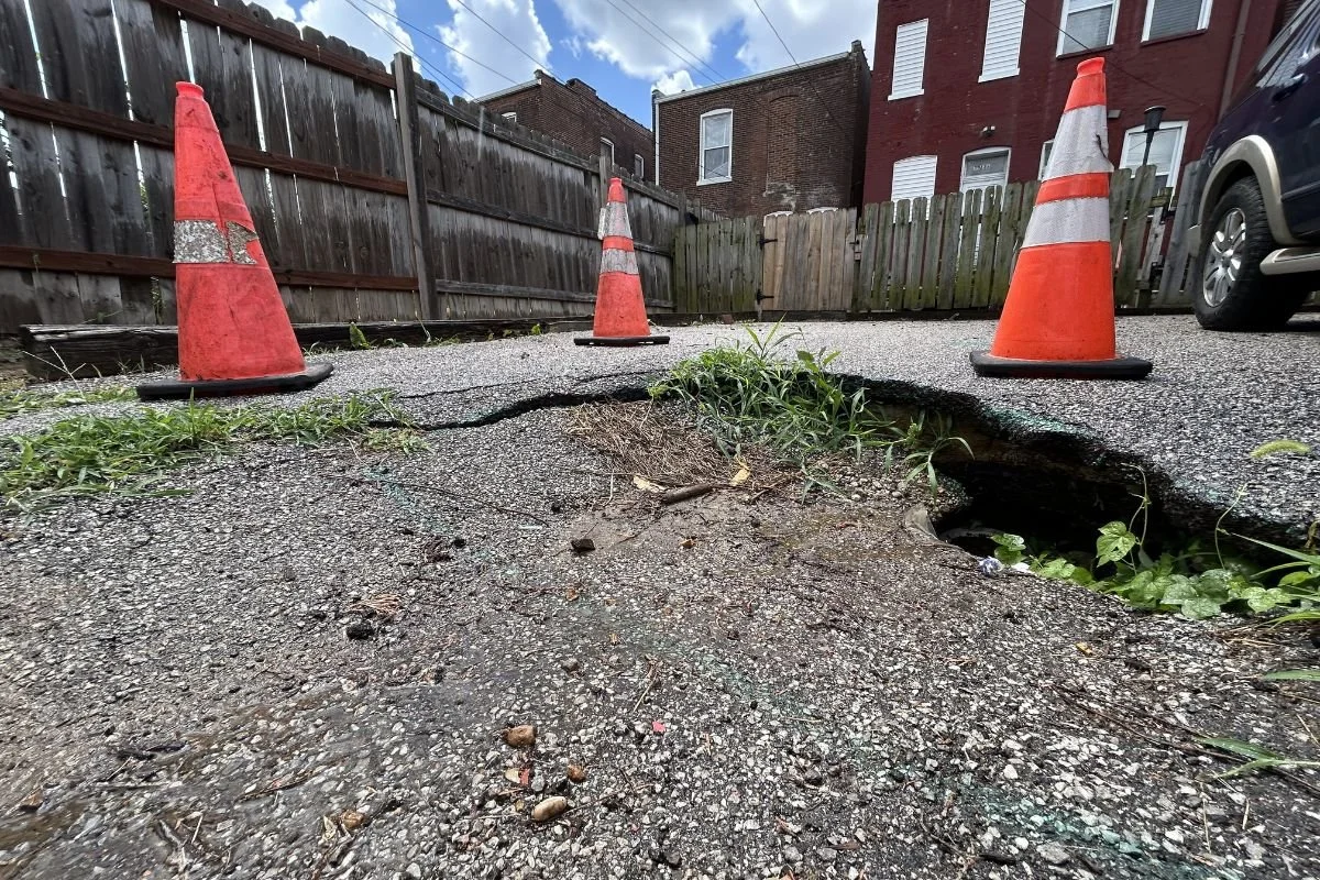 A sinkhole in a st. Louis city alley that has orange cones around the perimeter. you can see trash and pipes inside the deep hole.