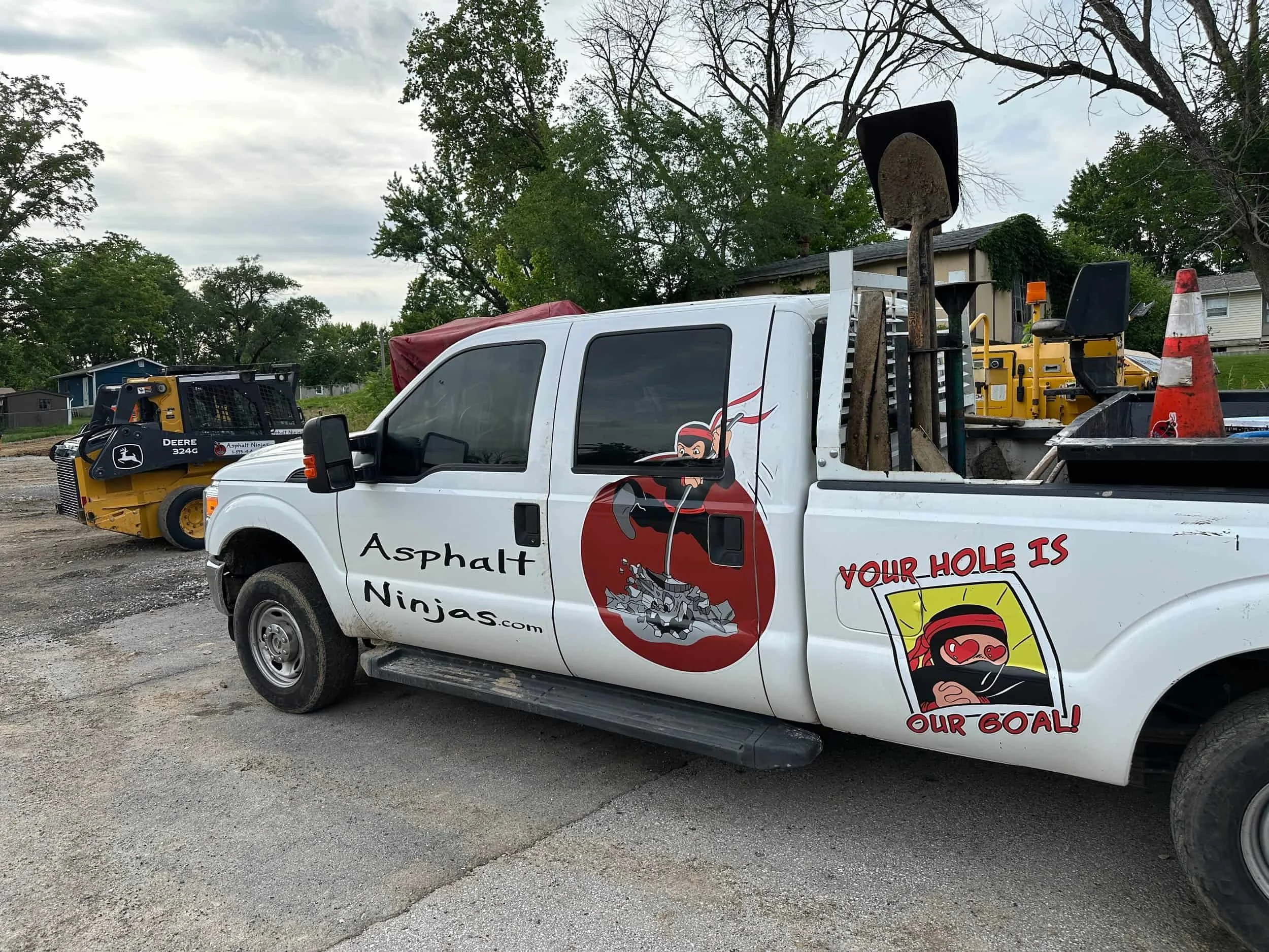 White pickup truck with Asphalt Ninjas logo, tools, and traffic cones in the bed, parked near construction equipment.