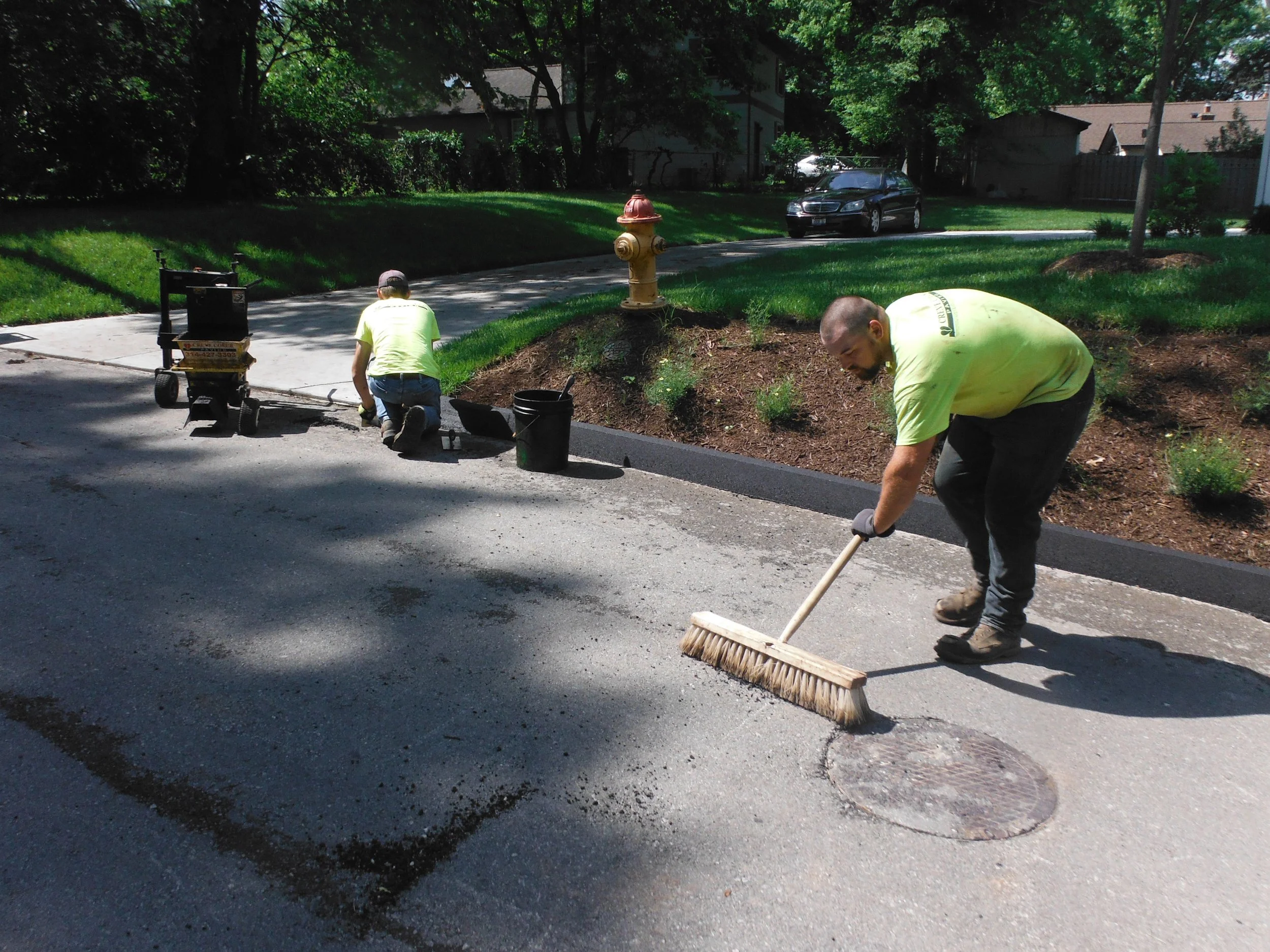 Two workers in yellow shirts repairing a pothole on a residential street, with one sweeping and the other kneeling near the curb.