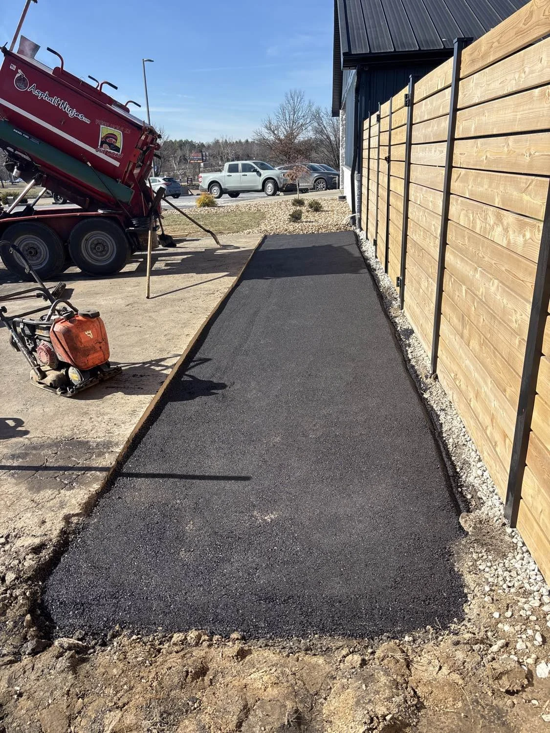 Freshly laid asphalt pathway beside a wooden fence, with construction equipment and a truck nearby on a sunny day.