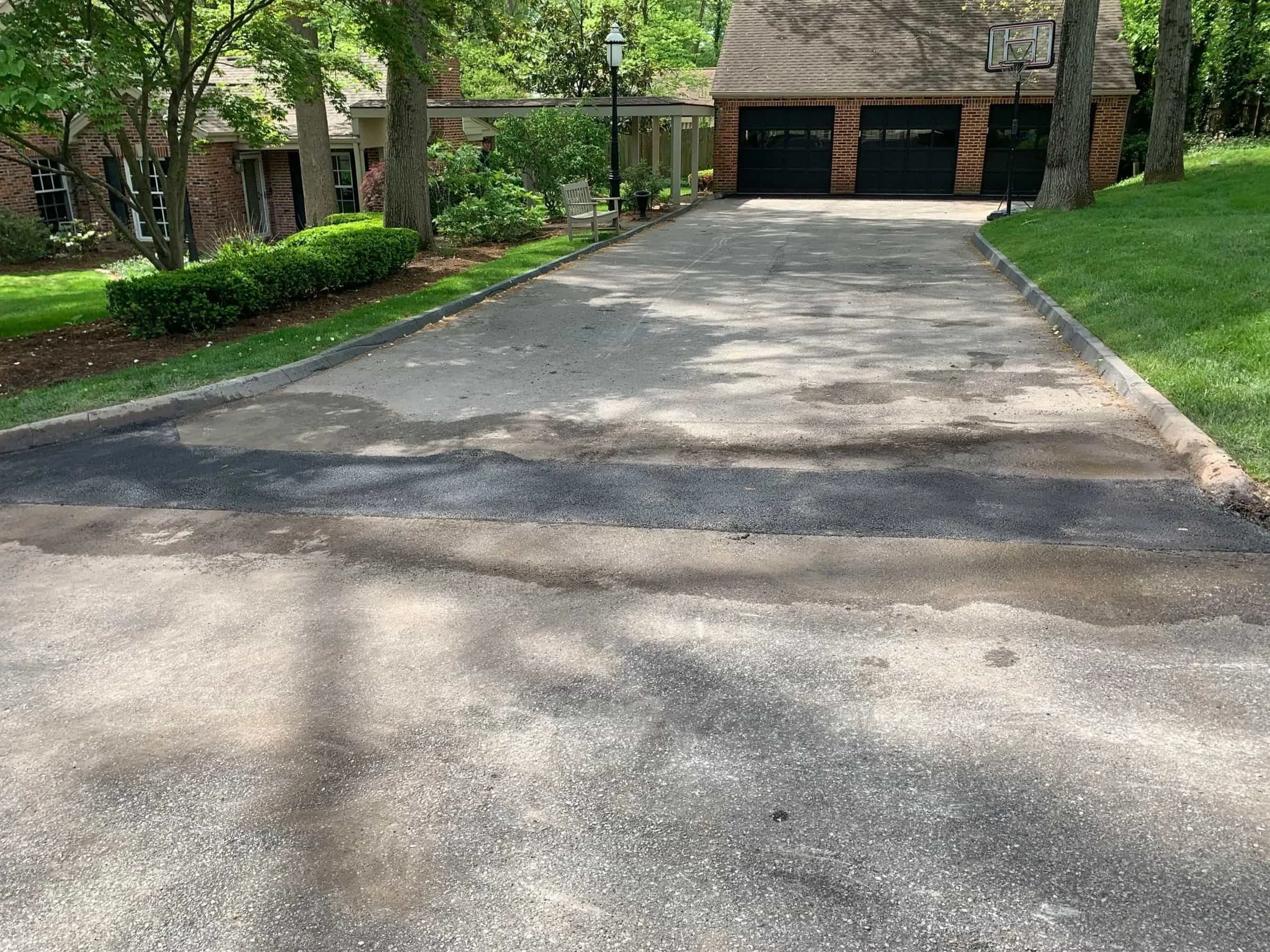 A paved driveway leads to a brick house with a basketball hoop above the garage doors, surrounded by trees and greenery.