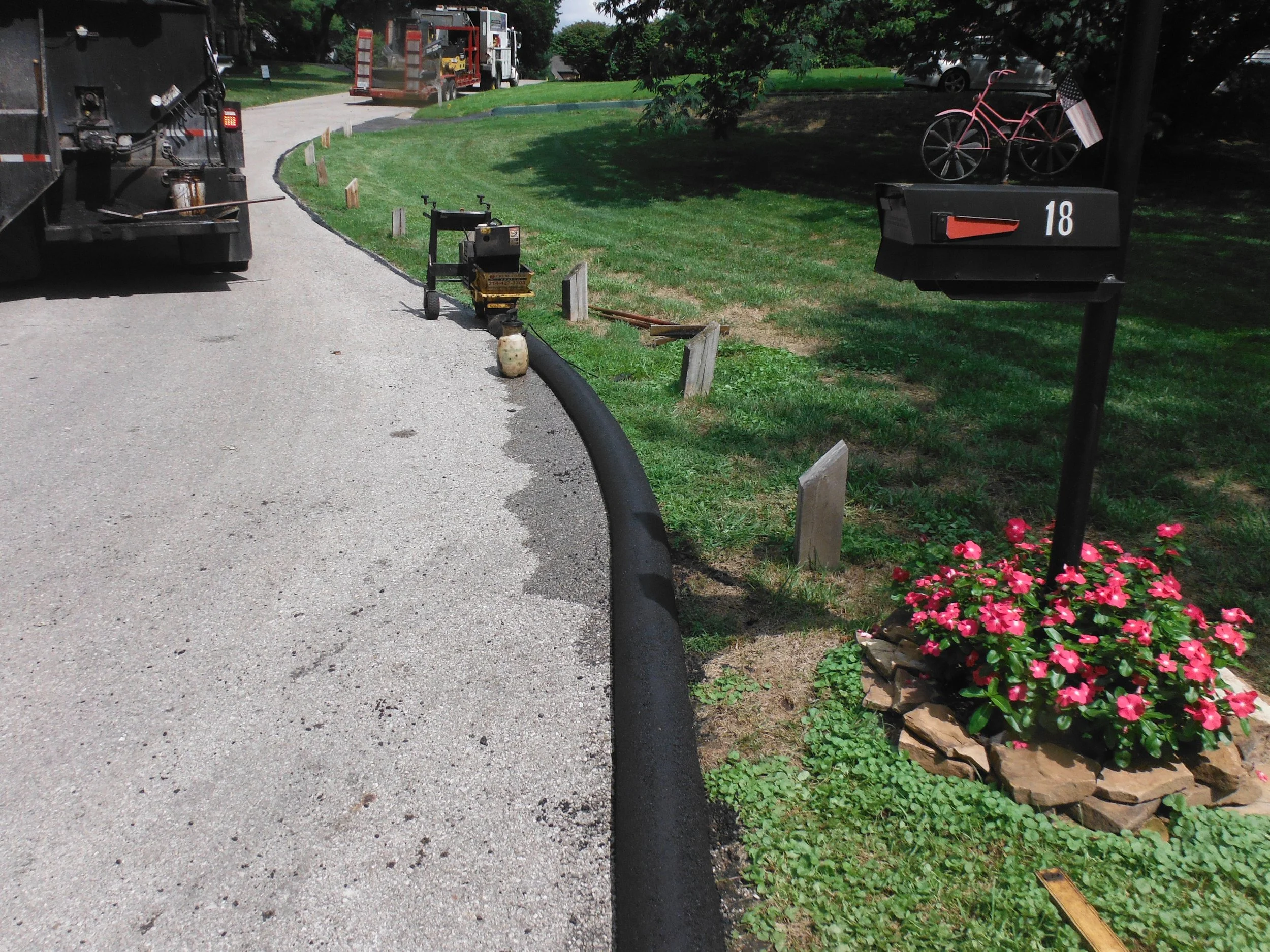 A street with a worker truck and equipment laying down new black asphalt along the side of the road. There is a small flower bed with pink flowers, a pink bicycle with an American flag, and a black mailbox marked with the number 18.