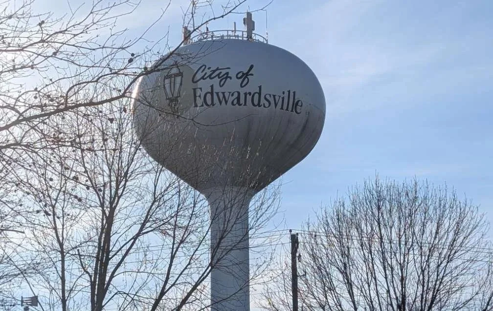 A water tower labeled City of Edwardsville stands among leafless trees under a clear blue sky.