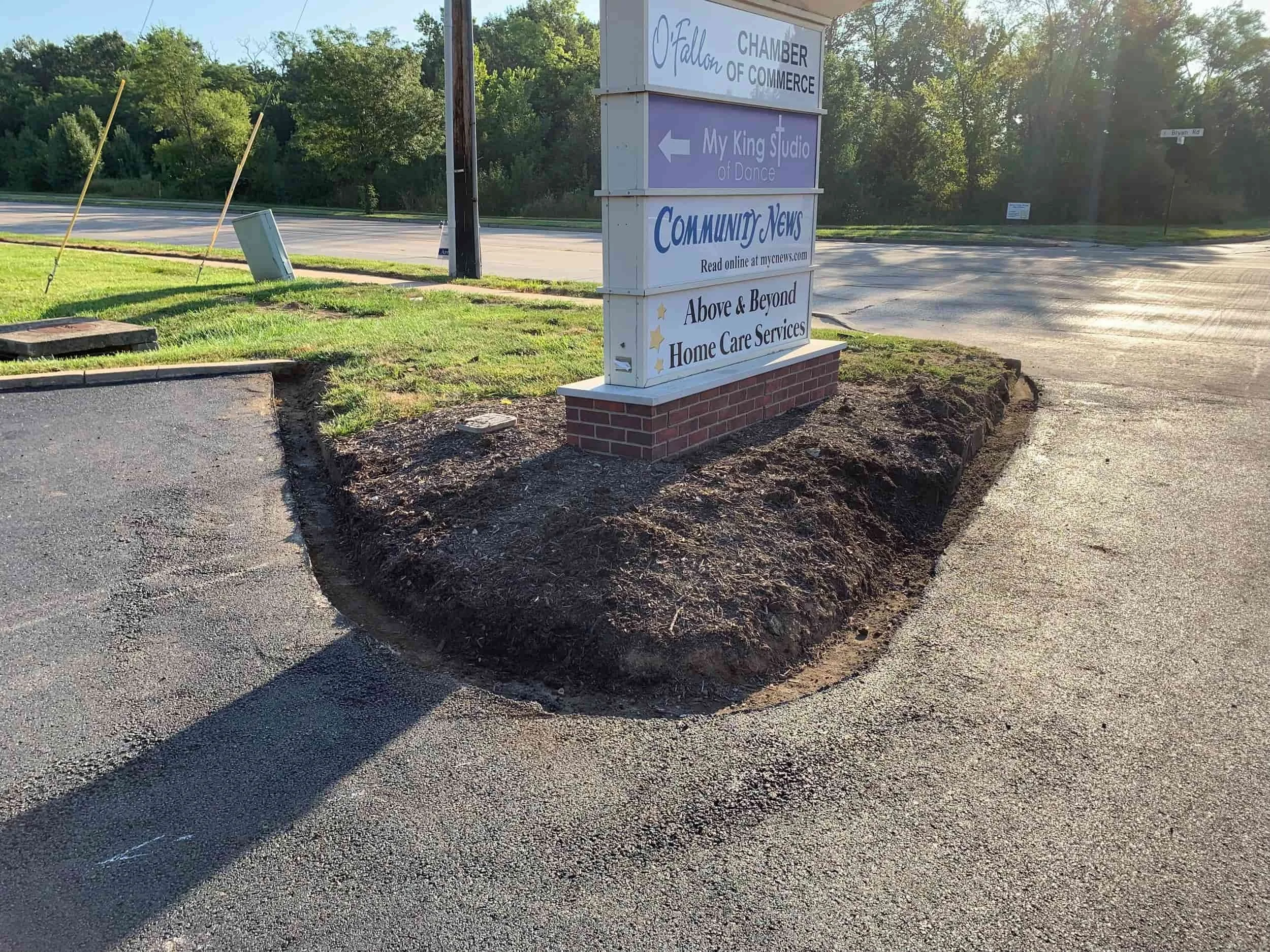 A patch of bare dirt surrounds a business sign next to a parking lot and an empty street on a sunny day.
