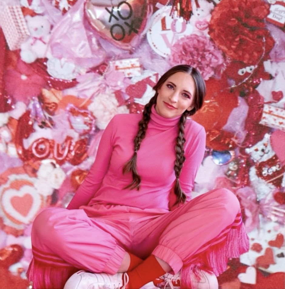 A woman with long braids, wearing a pink outfit, sitting cross-legged with a background filled with Valentine's Day themed decorations, including hearts, plush toys, and love-themed signs. The Valentine's Museum NYC Valentine's Day Event.