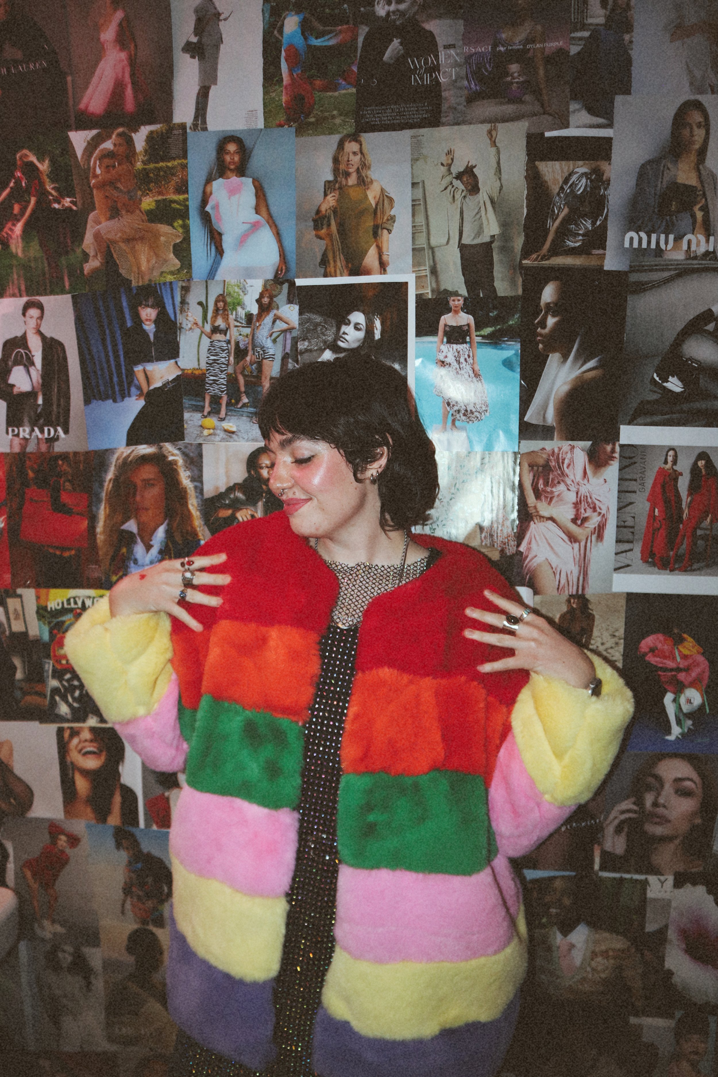 A young woman with short black hair and a septum piercing wearing a colorful, striped faux-fur jacket, standing in front of a collage of fashion and celebrity photos on a wall. The Valentine's Museum NYC Valentine's Day Event.