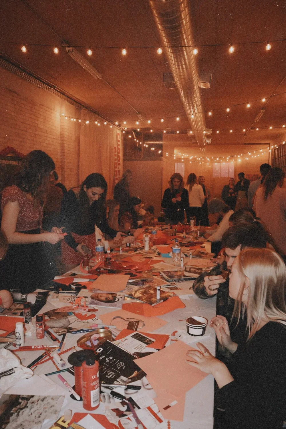 A group of women gathered around a cluttered table, engaging in a creative activity, in a warmly lit room with string lights and exposed brick walls. The Valentine's Museum NYC Valentine's Day Event.