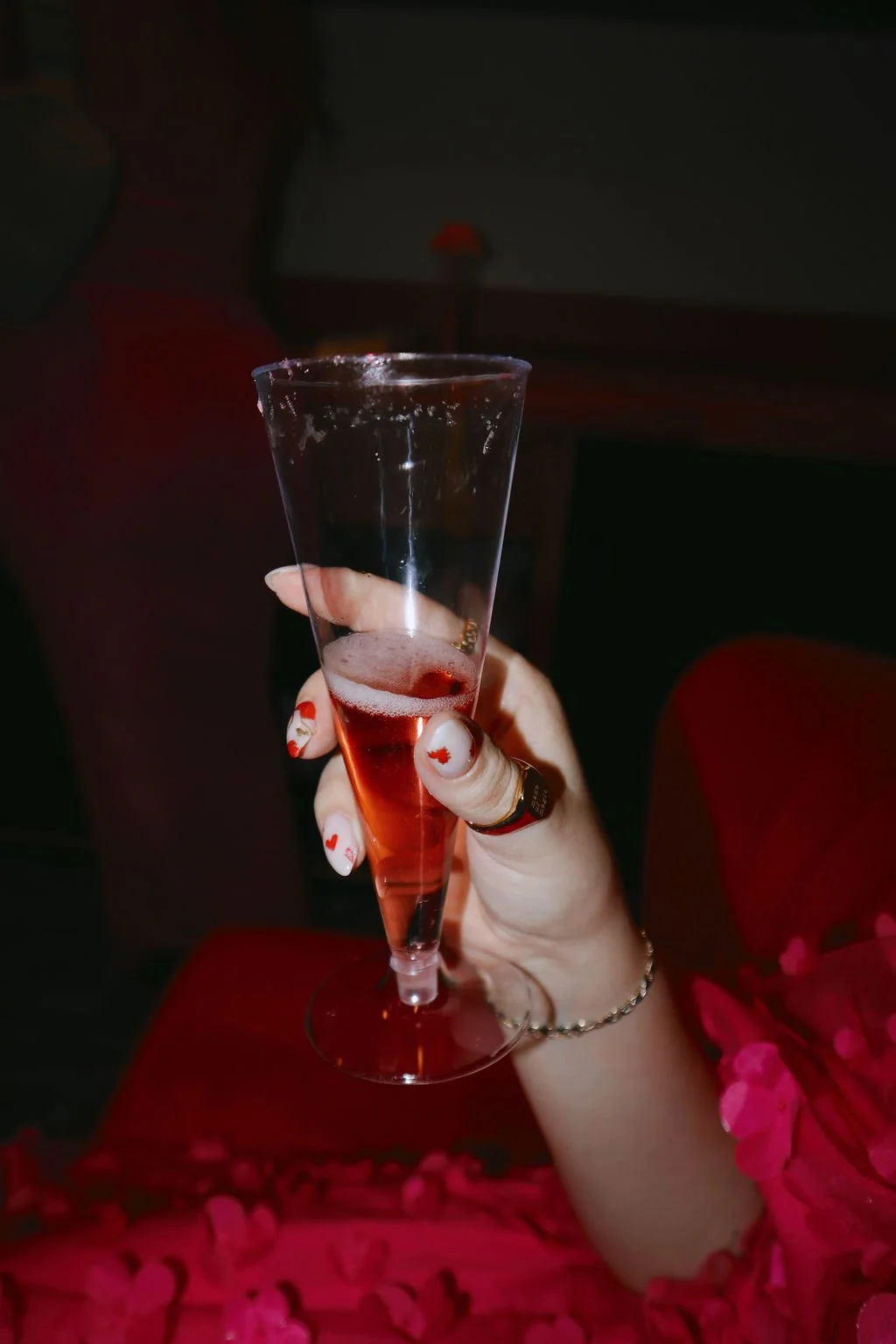 A person holding a champagne flute filled with pinkish sparkling wine or champagne, with decorative nail art and a ring on their finger, in a dimly lit setting. The Valentine's Museum NYC Valentine's Day Event.