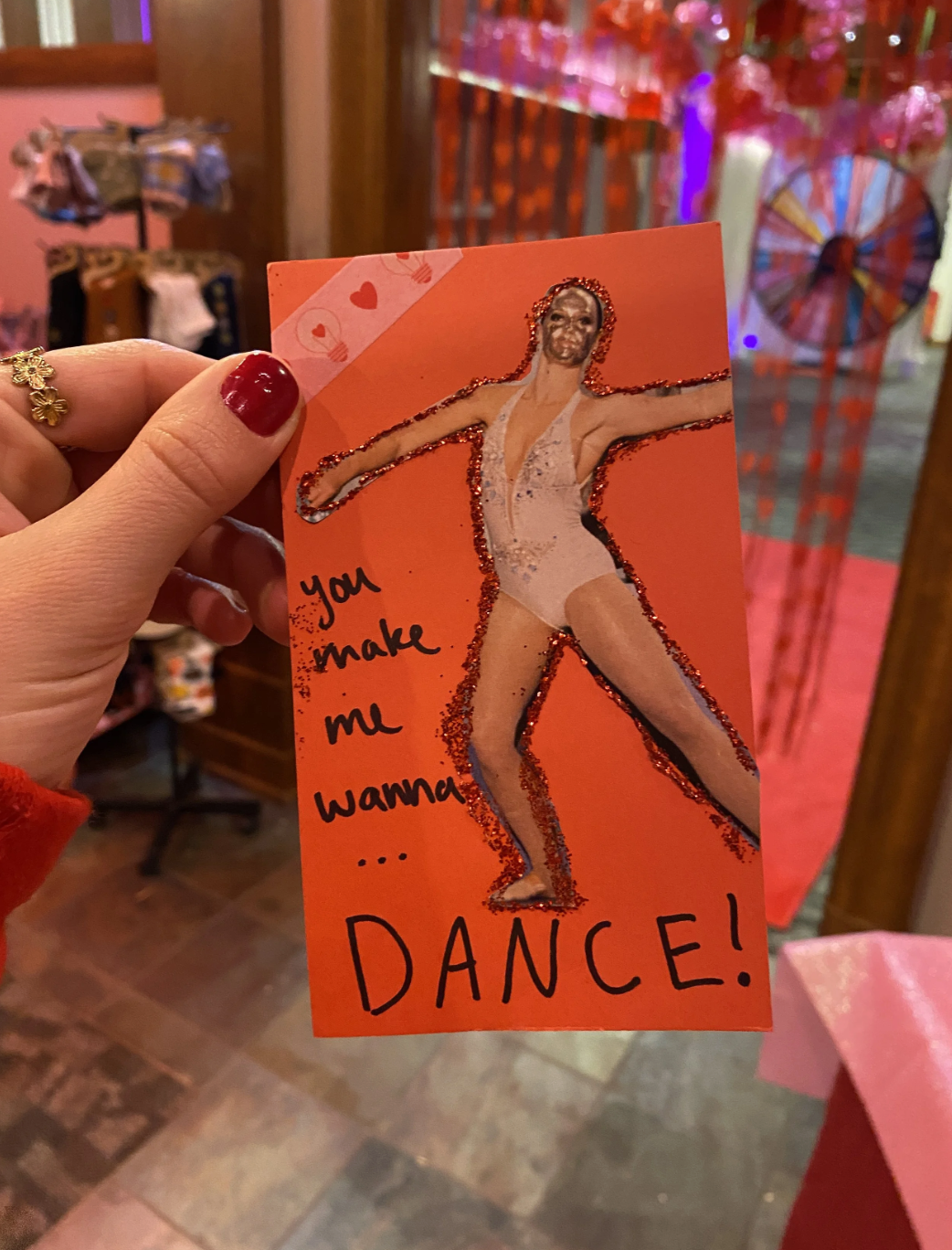 A handmade Valentine card featuring a glittery cut-out of a woman in a white leotard dance pose, with handwritten text that says 'You make me wanna... DANCE!' The card is held by a person with red nail polish, in a store with pink and red decorations in the background.