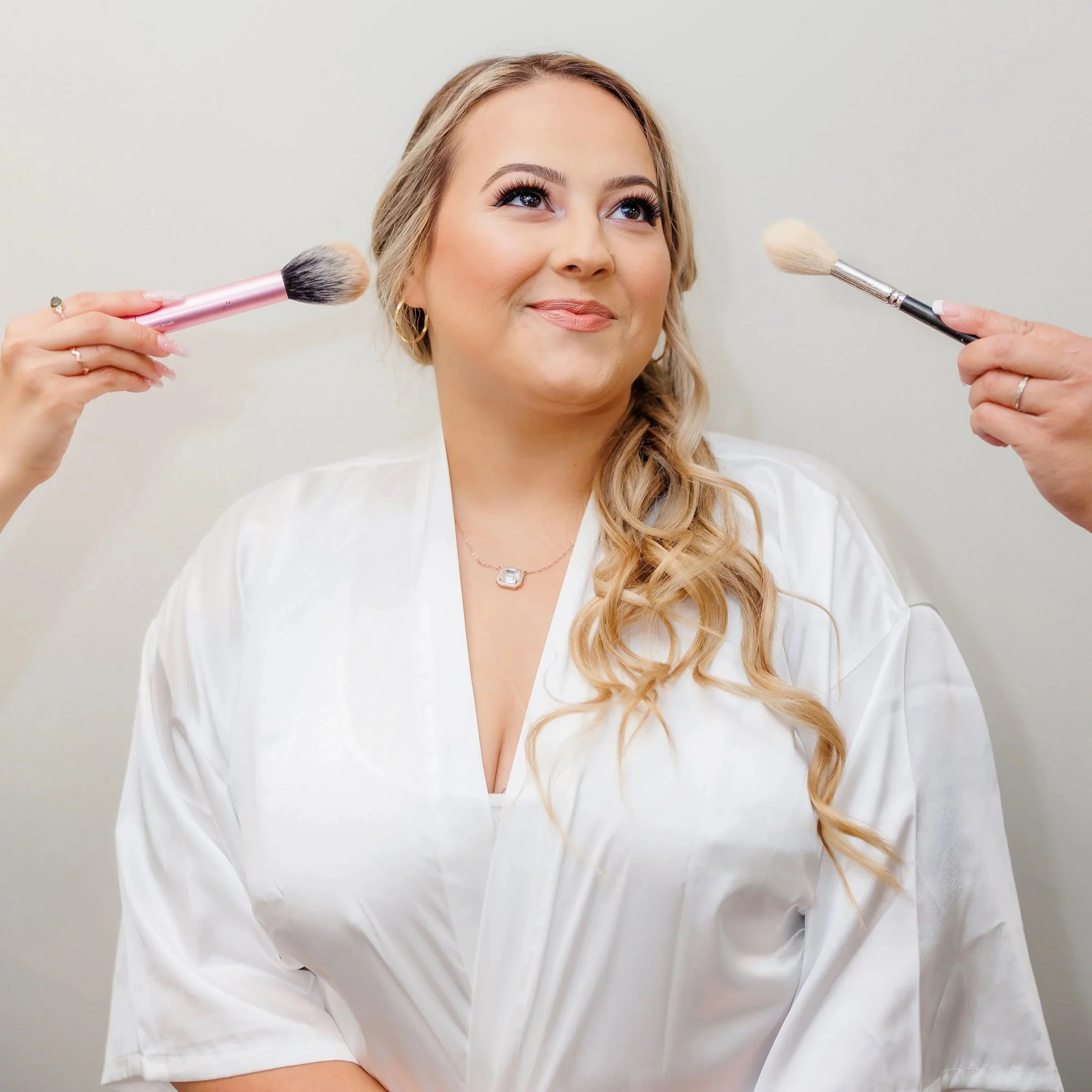 A woman with long, wavy blond hair and makeup, smiling slightly, being surrounded by two people holding makeup brushes, one on each side.