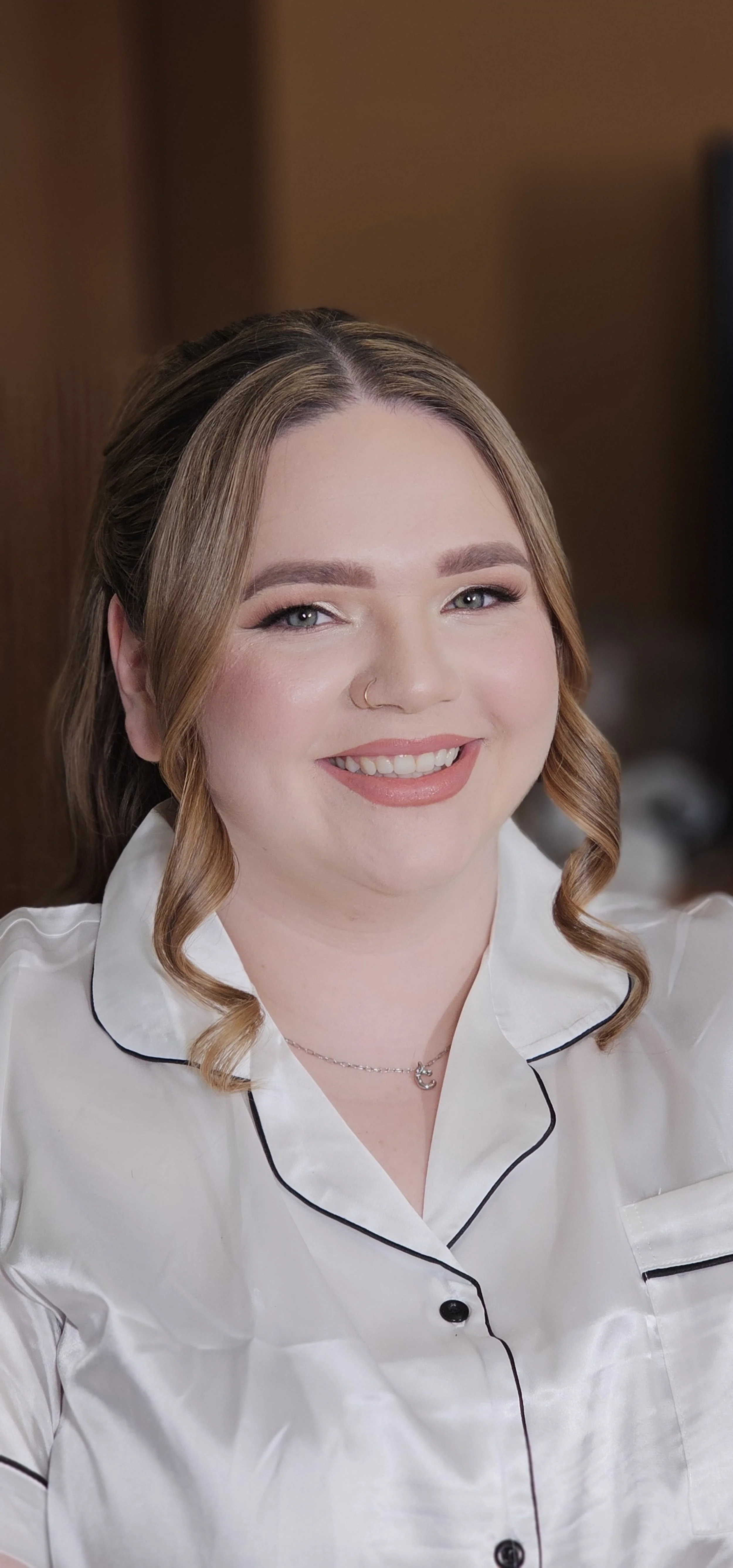Close-up of a smiling woman with light skin, blonde hair styled in loose curls, wearing a white satin pajama top with black piping, a silver necklace, and a nose ring.