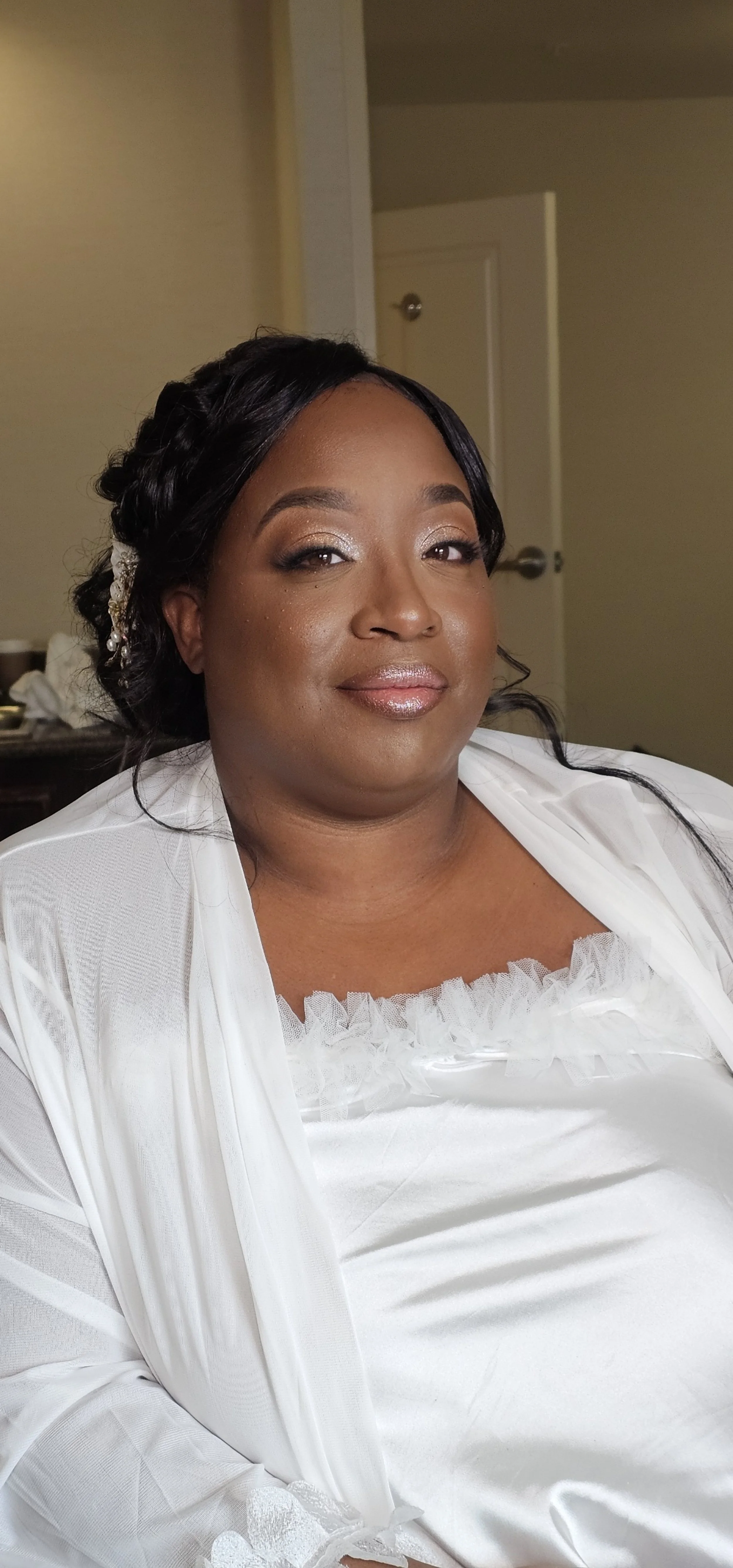 Close-up of a woman with dark hair styled in waves and a white satin and lace dress, sitting in a room with beige walls.