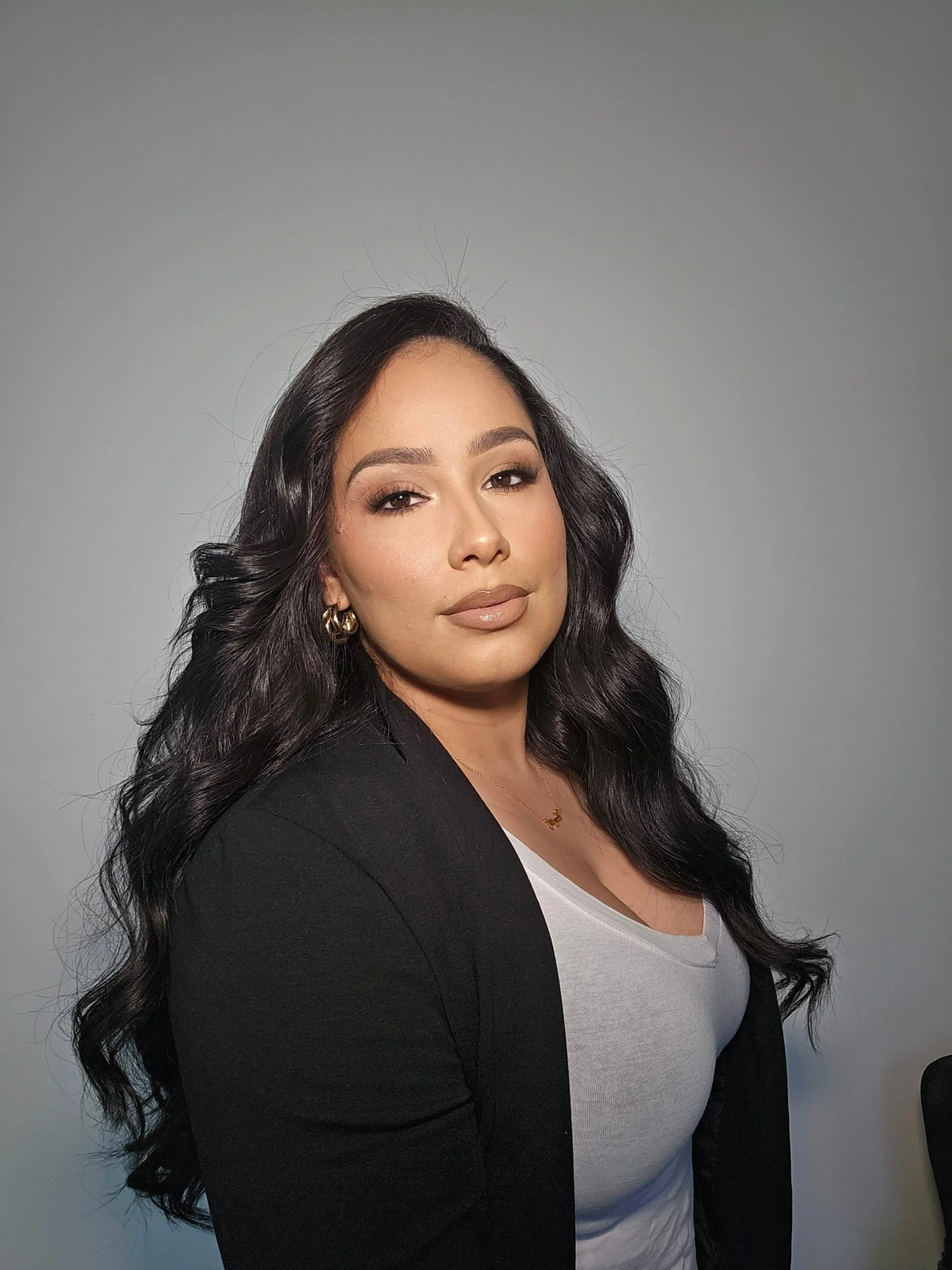 A woman with long dark hair wearing a black blazer and white top, looking at the camera against a plain gray background.