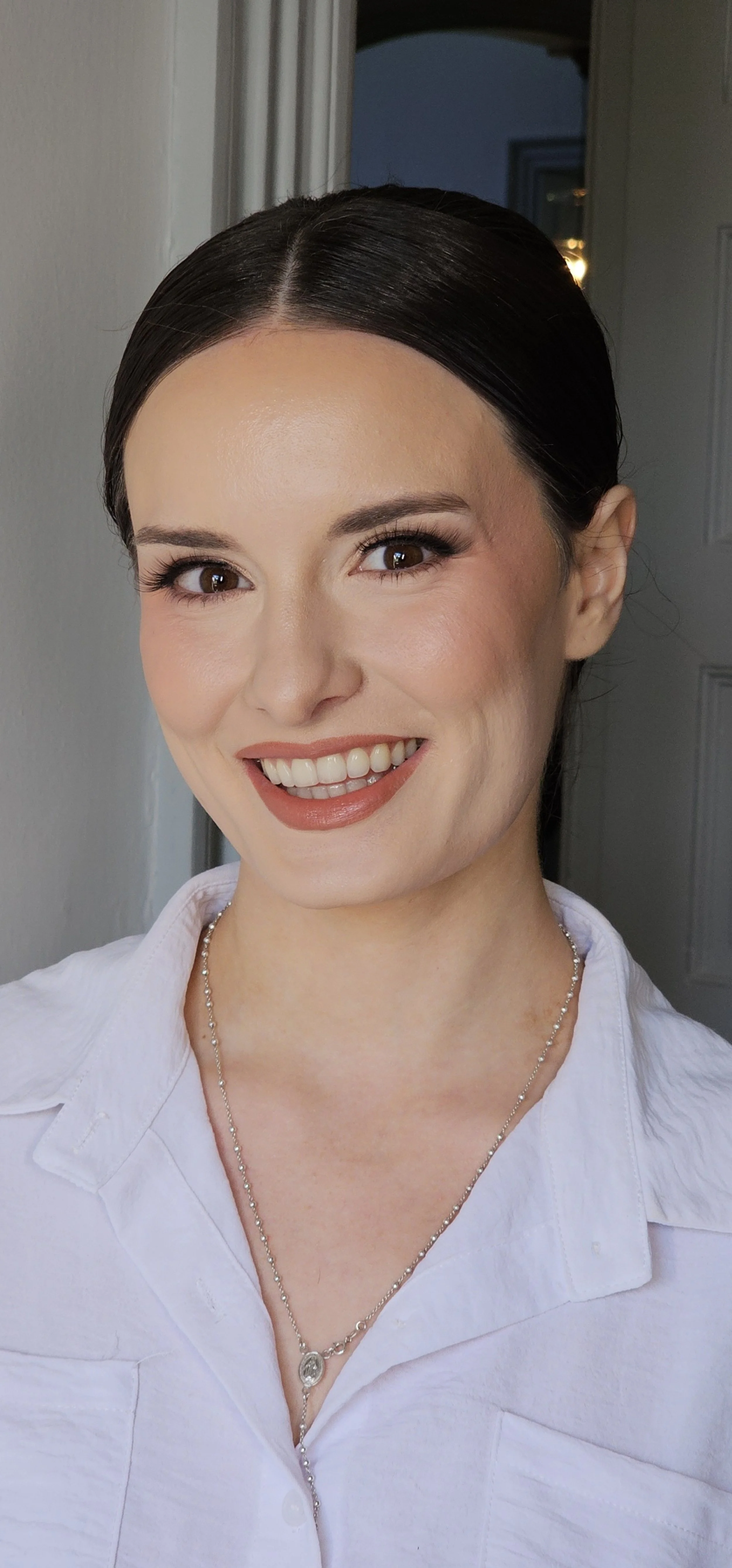 A woman with dark hair styled in a sleek bun, wearing a white shirt and a silver necklace, smiling at the camera indoors.