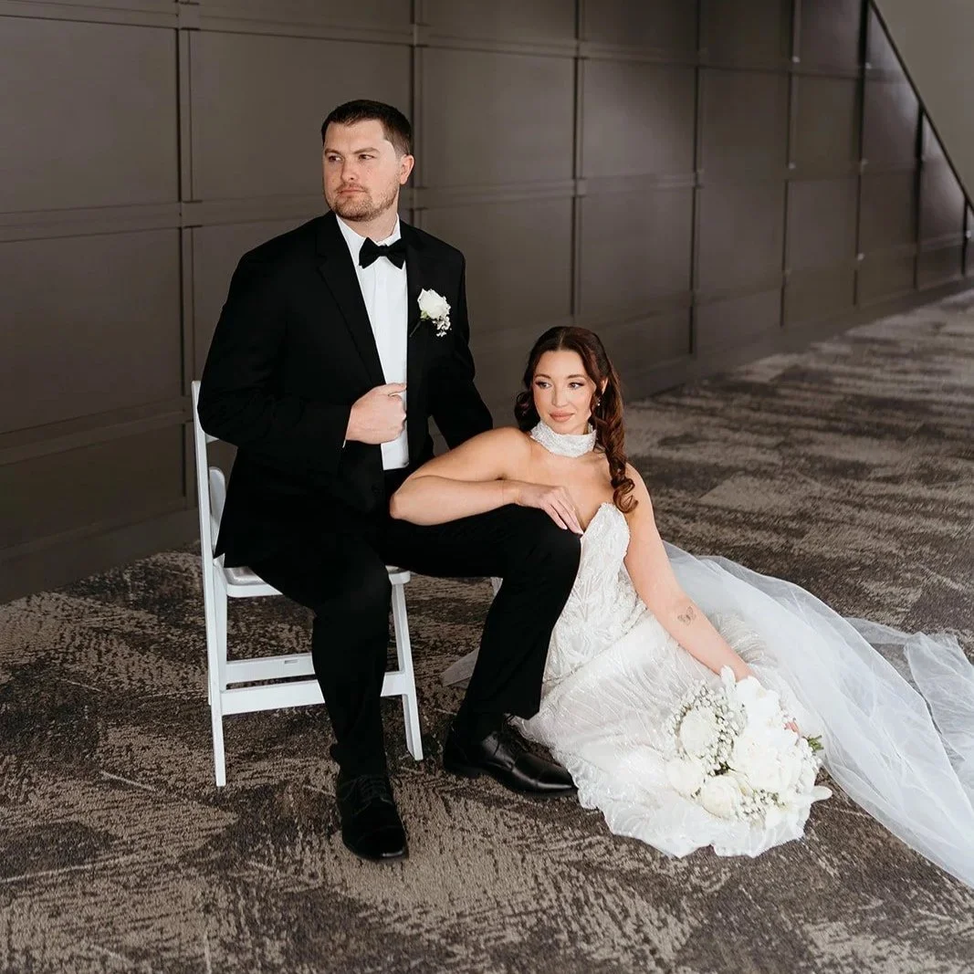 A bride and groom pose for a wedding photo indoors, with the groom seated on a chair and the bride sitting on the floor beside him, holding a bouquet of white flowers, both dressed in formal wedding attire.