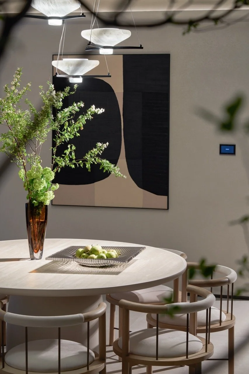 A modern dining room with an oval white table, surrounded by six light wood chairs with white cushions. A tall brown vase with green and white flowers is on the table, along with a silver tray of green apples. Above, three contemporary pendant lights hang from the ceiling. A black and beige abstract art piece is on the wall in the background.