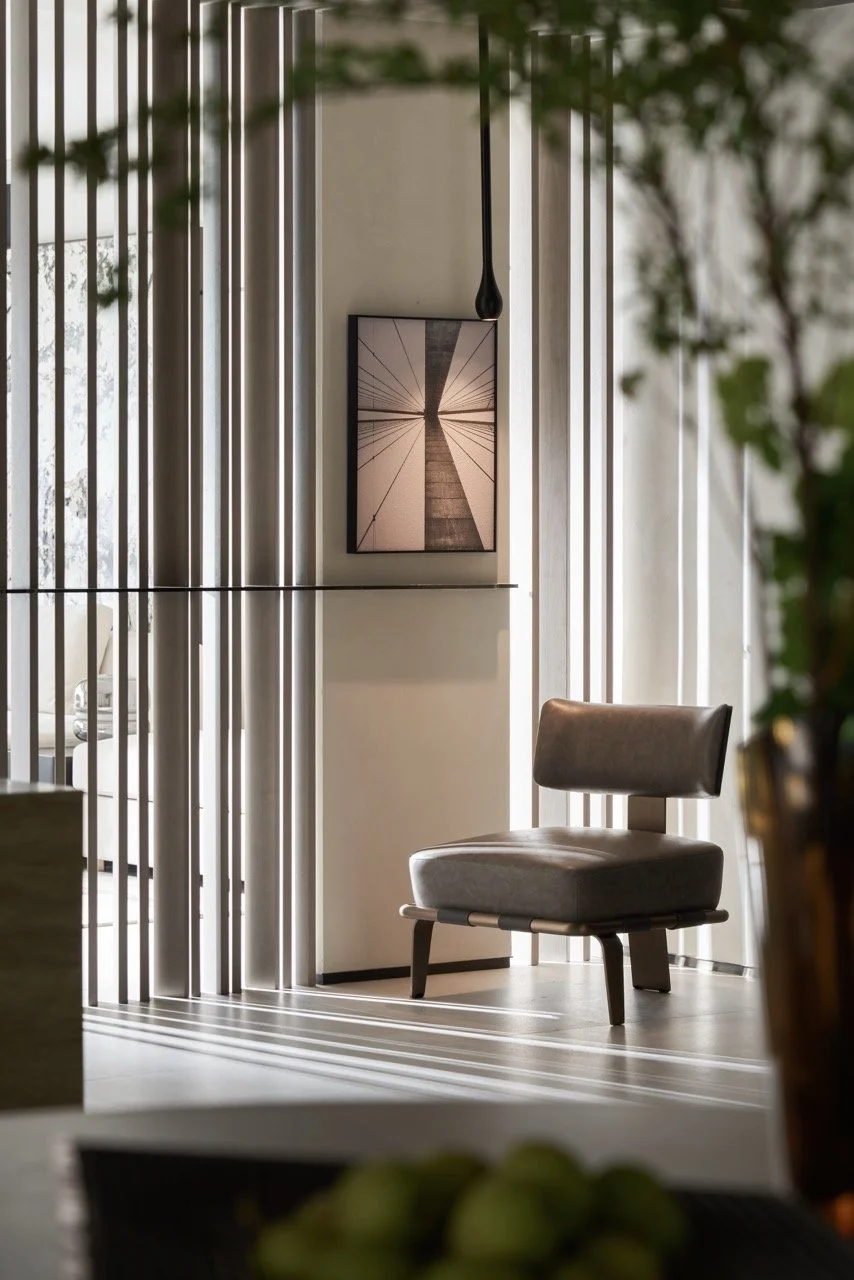 Interior of a modern room with sunlight creating striped shadows on the floor, a gray lounge chair, a minimalist picture on the wall, and vertical white slats acting as a room divider.