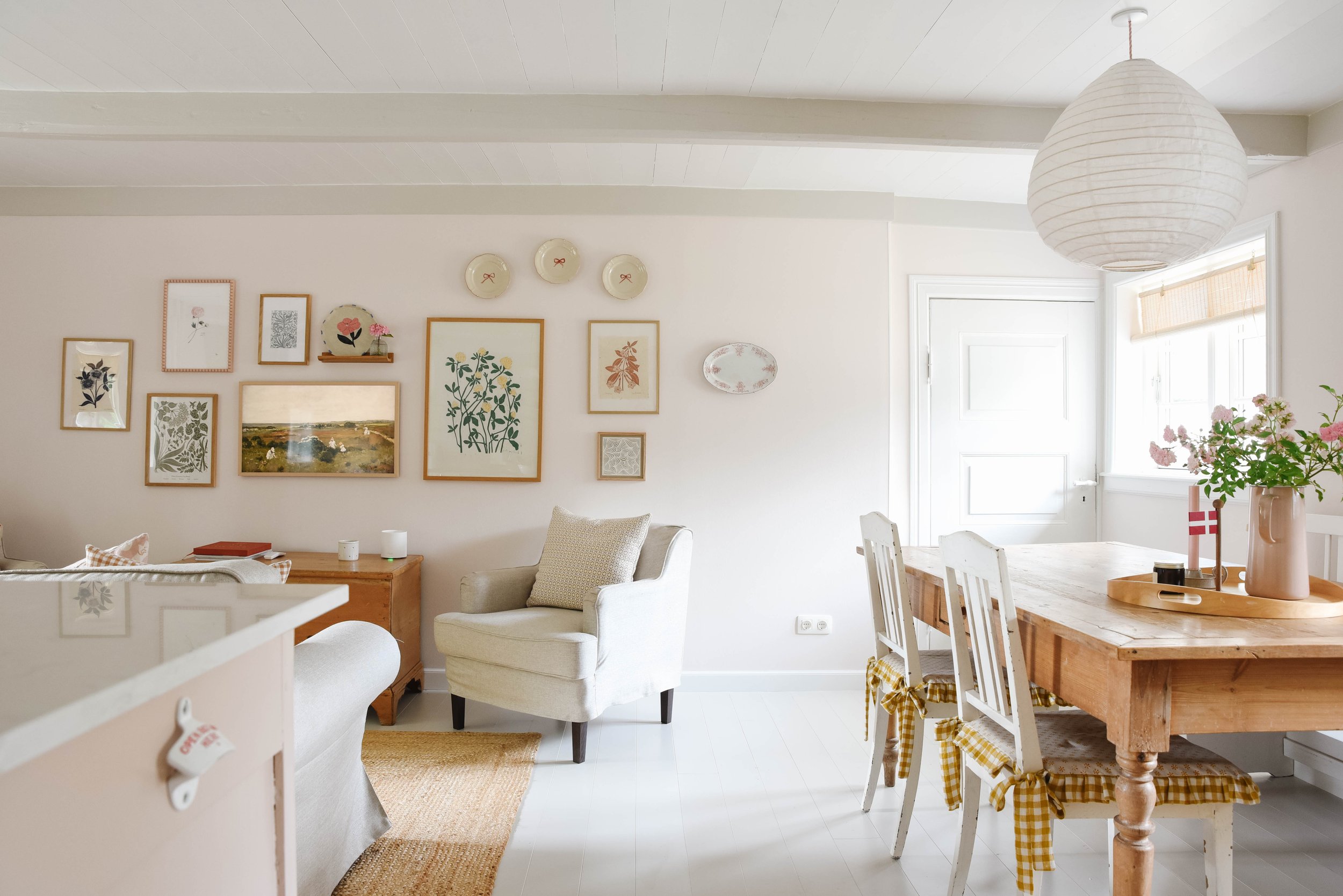 A cozy, light-filled dining and living room area decorated with framed botanical art and decorative plates on the wall, a large paper lantern hanging from the ceiling, a wooden dining table with white chairs and yellow checkered accents, and a vase o