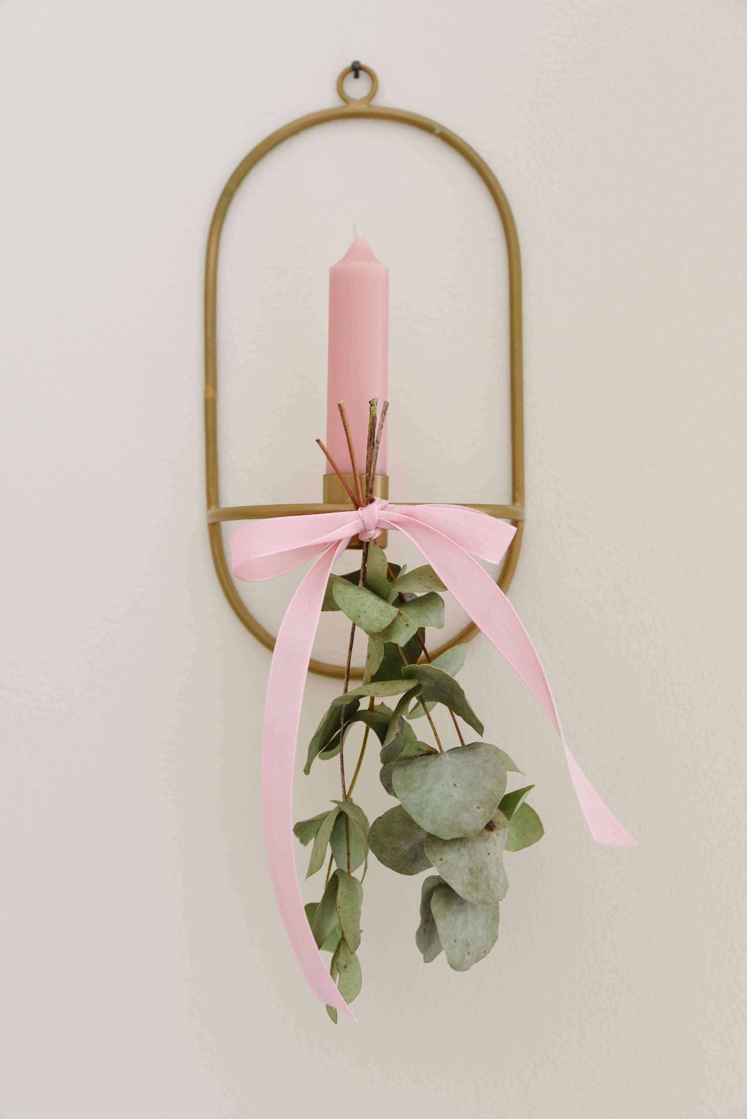 A pink candle with a white wick, tied with a pink ribbon and green leaves, hanging on a gold metal frame against a plain wall.