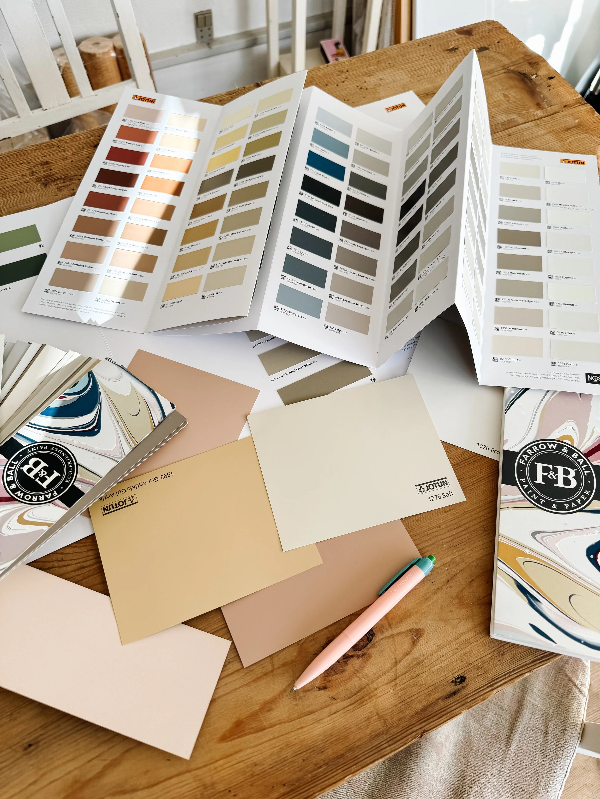 Color sample fan decks, paint color swatches, and paint samples spread out on a wooden table, with a pen and a small piece of paper nearby.
