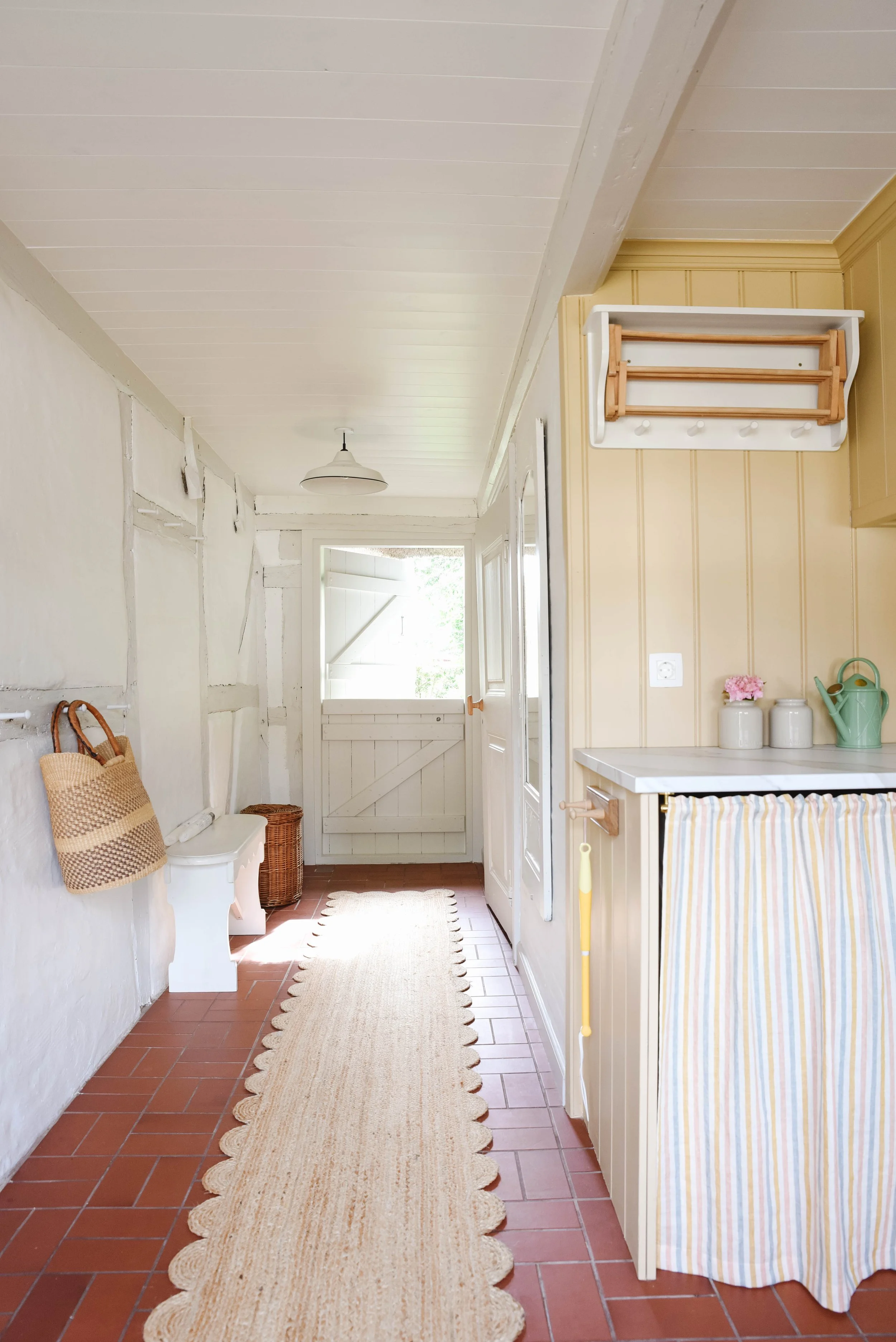 A cozy country kitchen with a beige striped curtain on the stove, white wall with yellow paneling, a beige rug on red brick flooring, and white ceiling with a hanging light fixture.