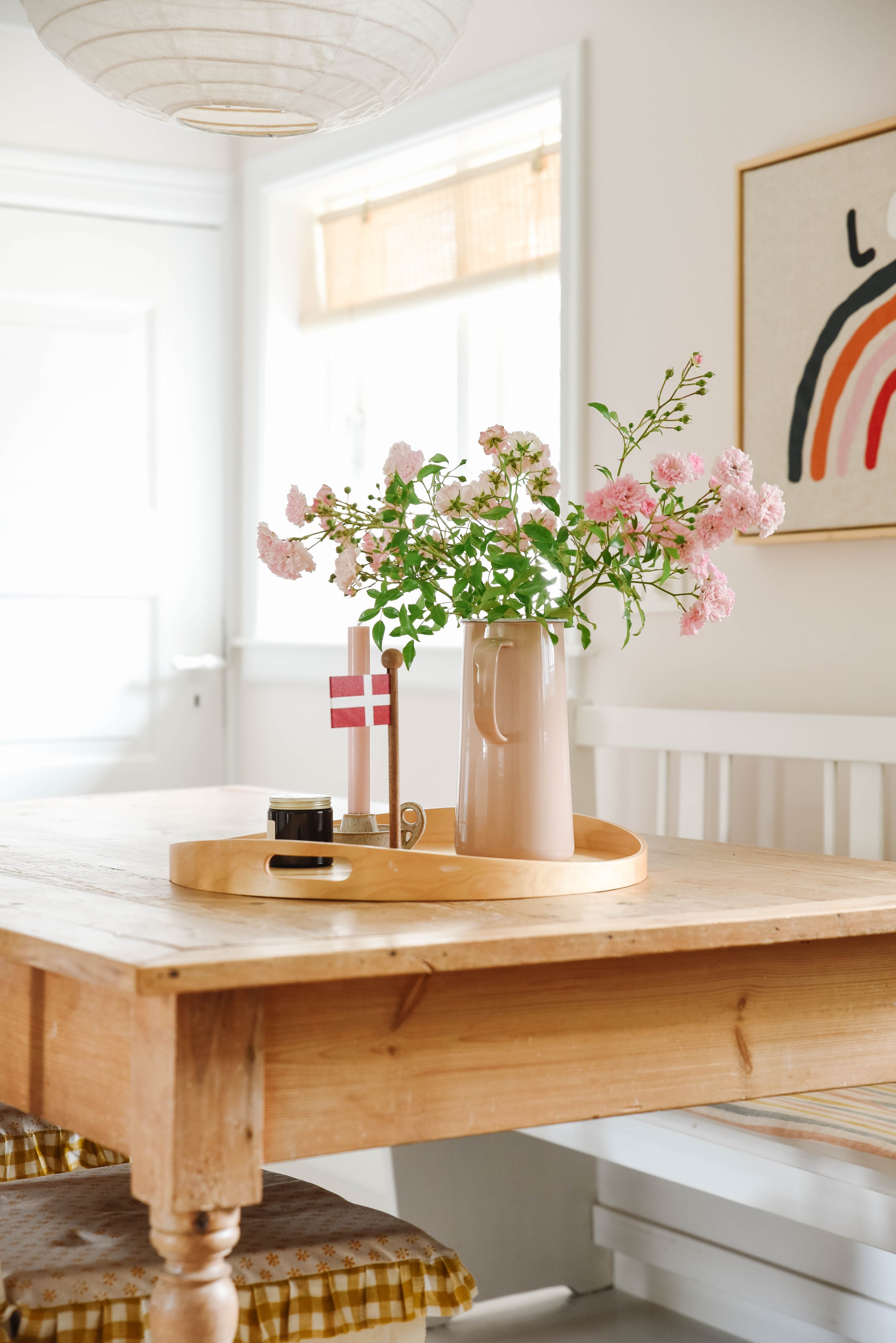 A wooden table with a tray holding a beige vase with pink flowers, a small Danish flag, and a small black container in a bright room with framed artwork and a window with a bamboo blind.