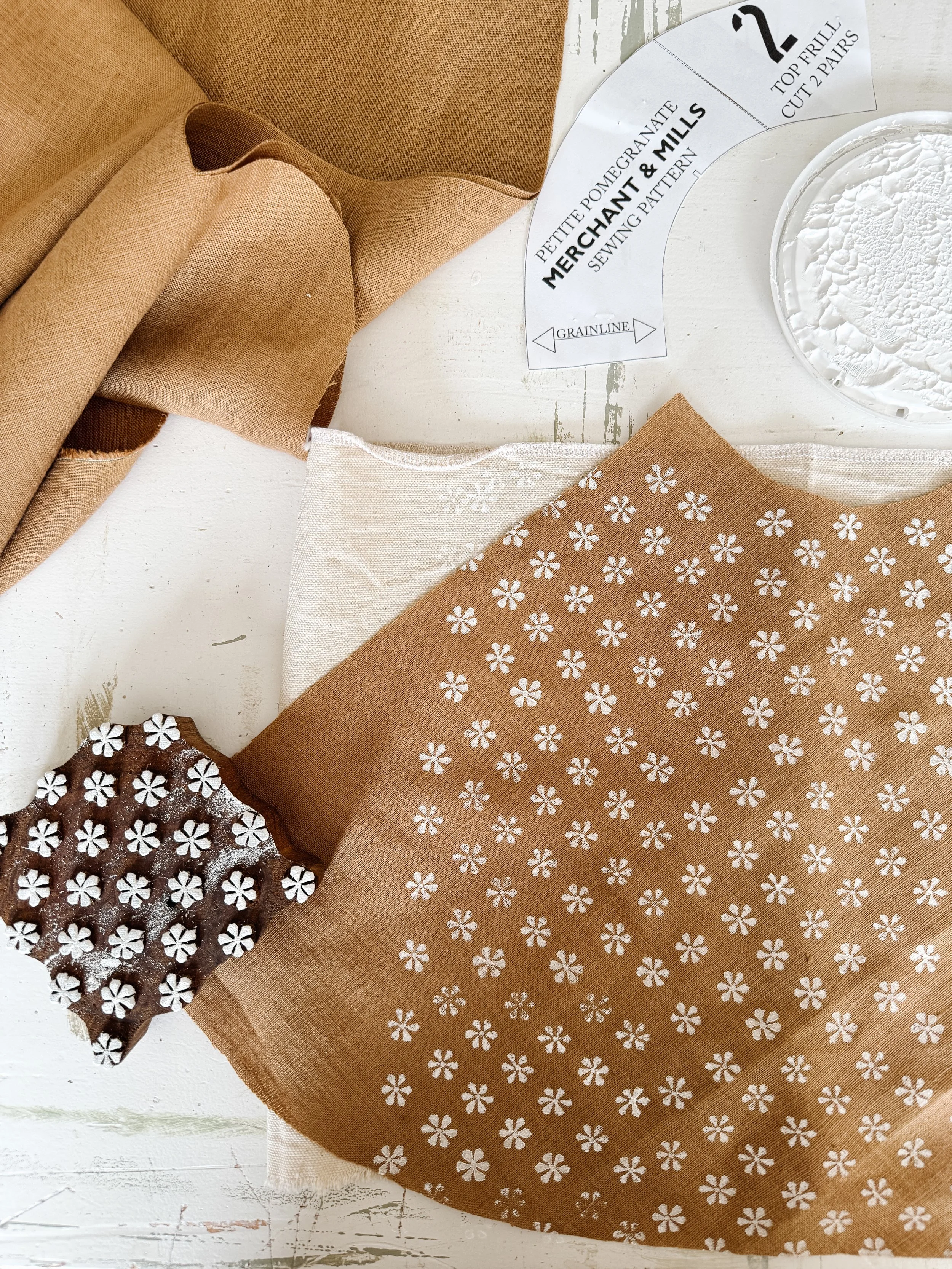 Various fabrics in shades of brown and beige, some with floral patterns, arranged on a white wooden surface, alongside sewing pattern pieces and a container of white powdered material.