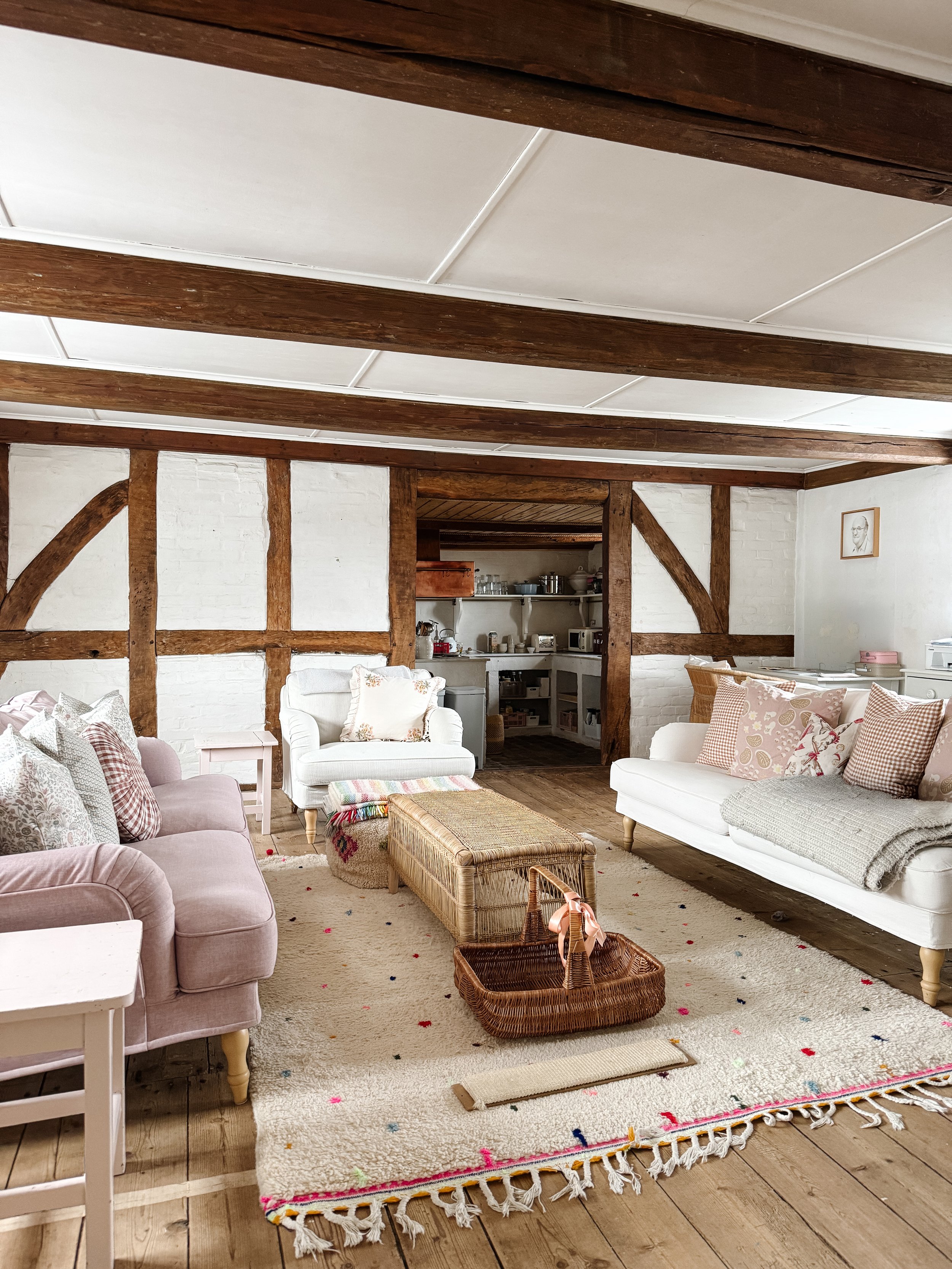 A cozy living room with pastel-colored sofas, a woven basket coffee table, a plush area rug with colorful specks, and wooden beams on the ceiling. A small wicker basket with a pink ribbon is on the floor, and a picture hangs on the white brick wall.