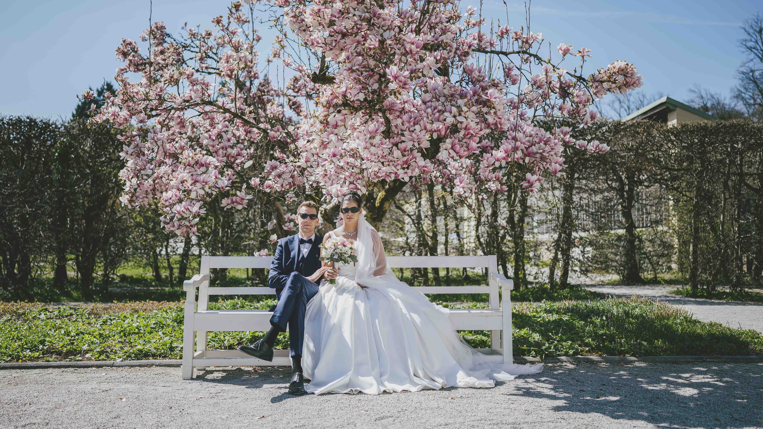 Ein Brautpaar sitzt auf einer weißen Bank unter einem blühenden Magnolienbaum in einem Garten, bei sonnigem Wetter, im Hintergrund Bäume und ein kleiner Weg.