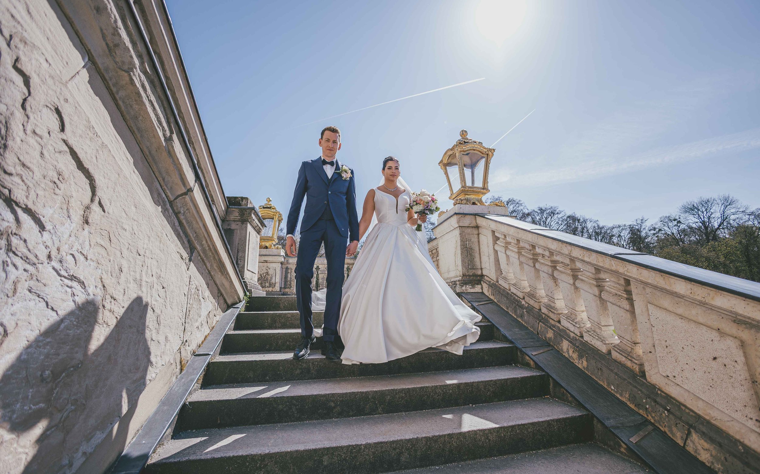 Hochzeitspaar auf der Treppe im Freien bei sonnigem Wetter, die Braut trägt ein langes weißes Brautkleid und hält einen Blumenstrauß, der Bräutigam trägt einen dunklen Anzug mit Fliege.