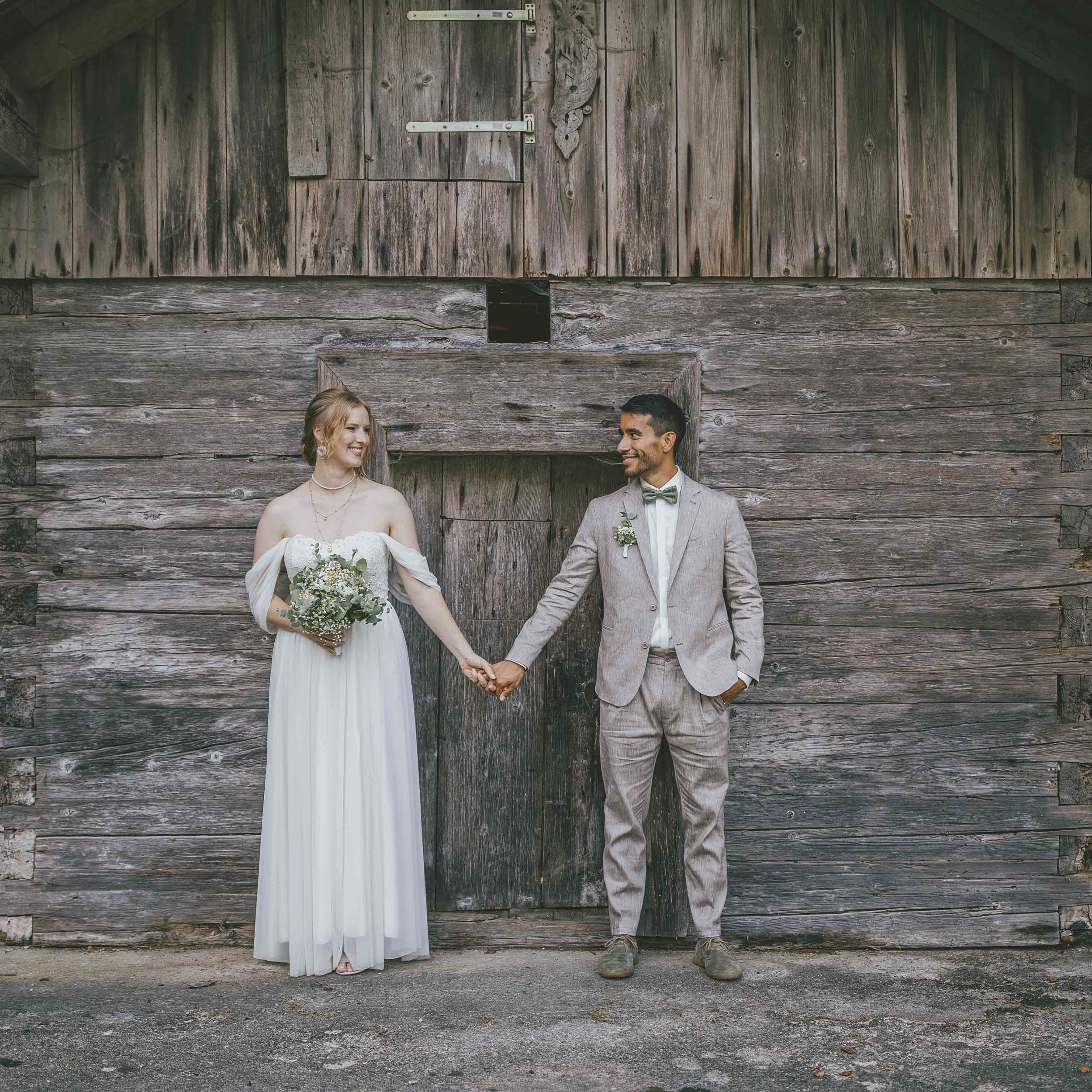 Ein Brautpaar hält Händchen vor einer alten Holzwand bei ihrer Hochzeit.