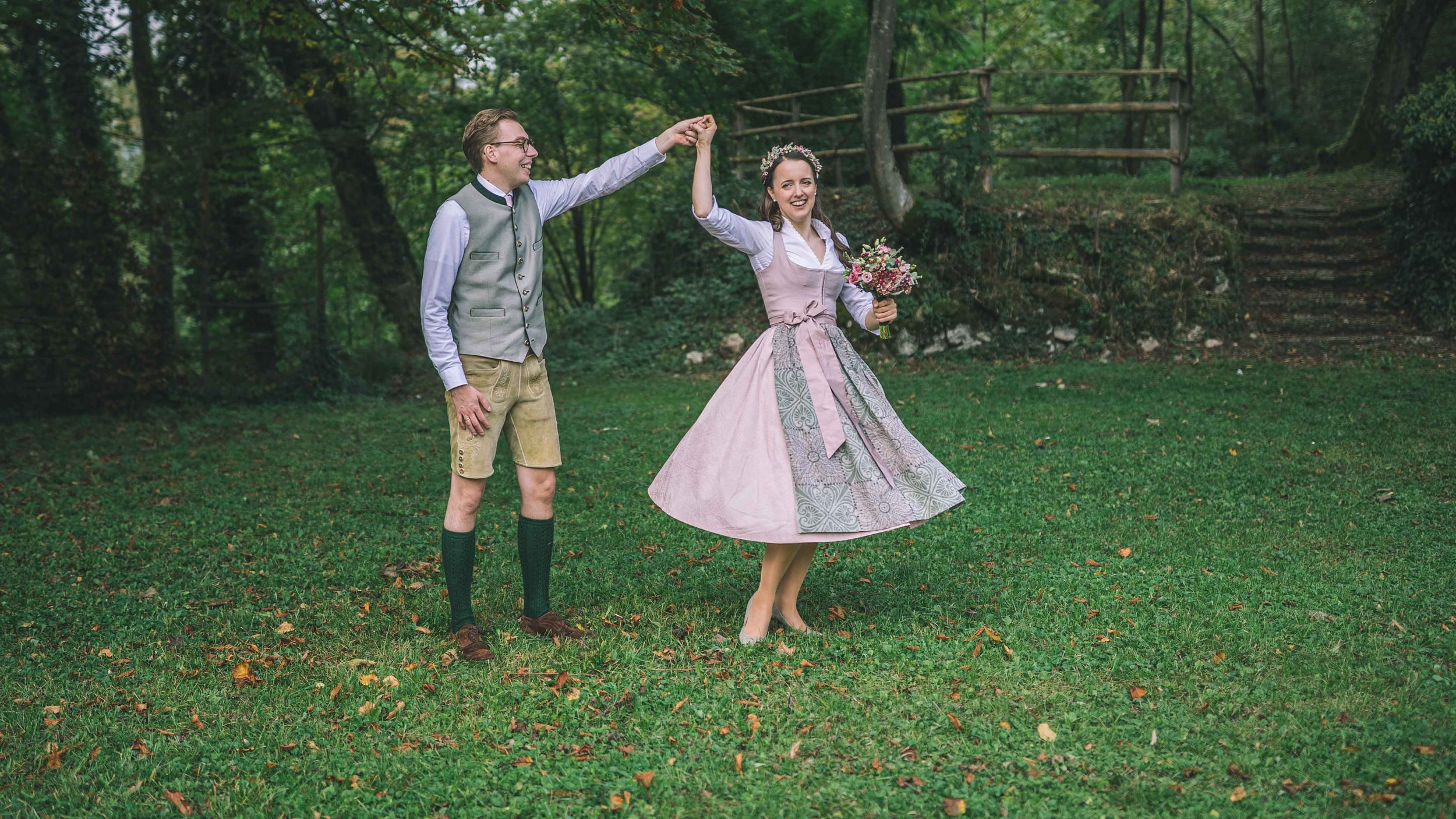 Ein Mann und eine Frau tanzen im Garten, der Mann trägt traditionelleKleider, hält die Hand der Frau, die ein Dirndl trägt und eine Blumenbouquet hält.