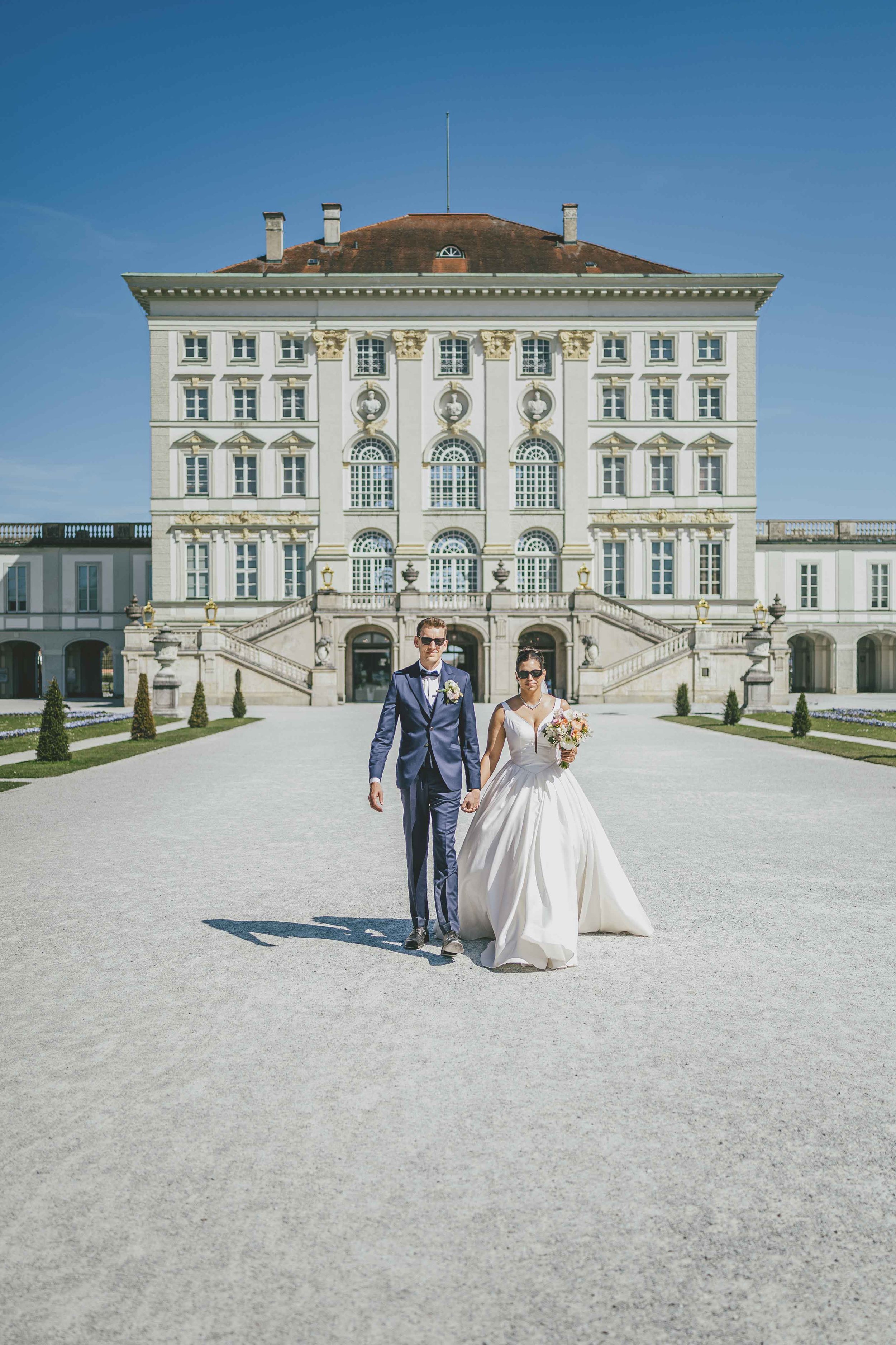 Braut und Bräutigam gehen vor einem großen, weißen Schloss an einem sonnigen Tag, die Braut trägt ein weißes Hochzeitskleid und hält einen Blumenstrauß, der Bräutigam trägt einen dunklen Anzug mit Fliege.