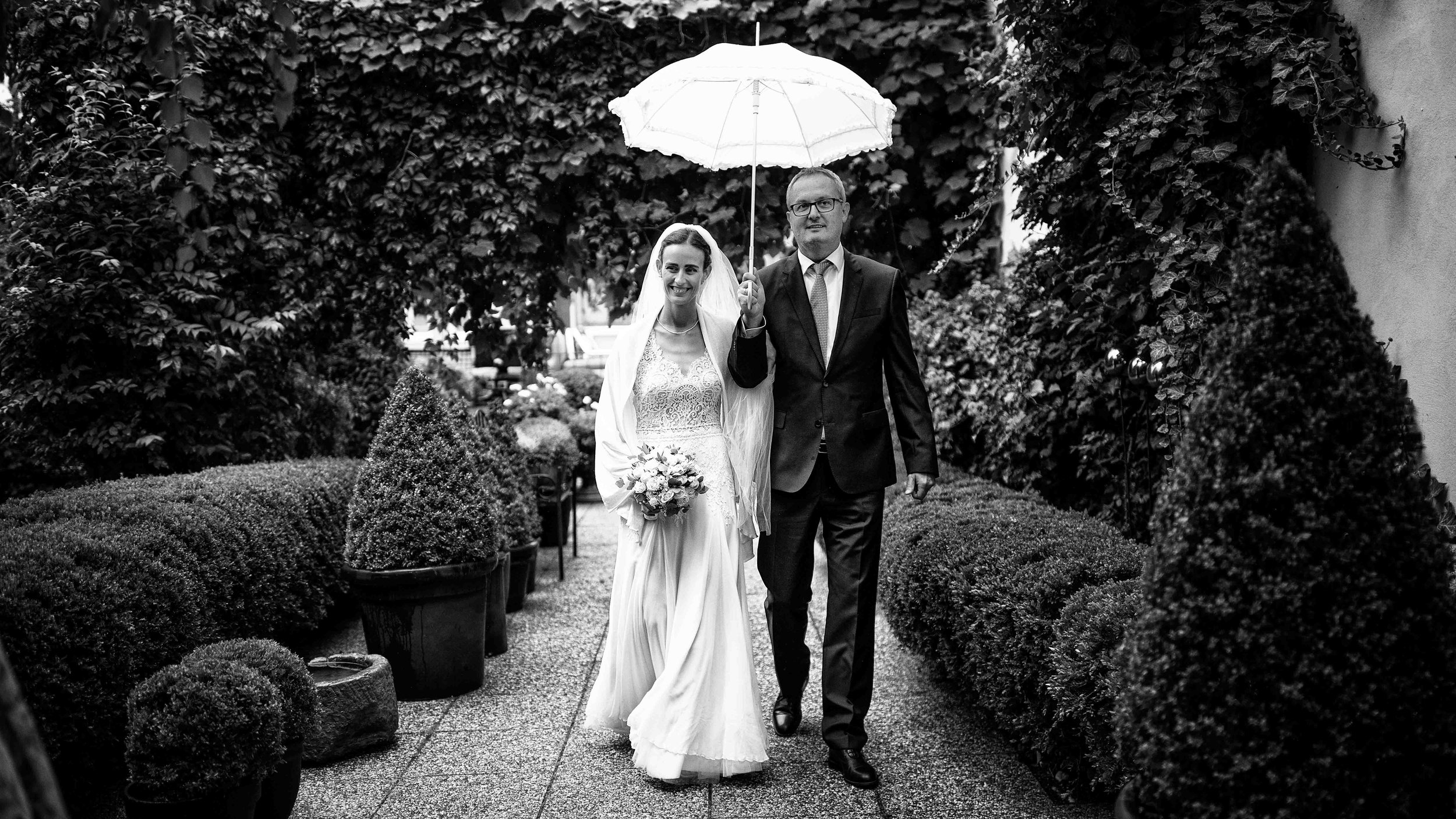 Ein glückliches Brautpaar spaziert an einem Regentag in einem Garten, wobei die Braut mit einem Blumenstrauß in der Hand neben dem Bräutigam geht, der einen weißen Regenschirm hält.