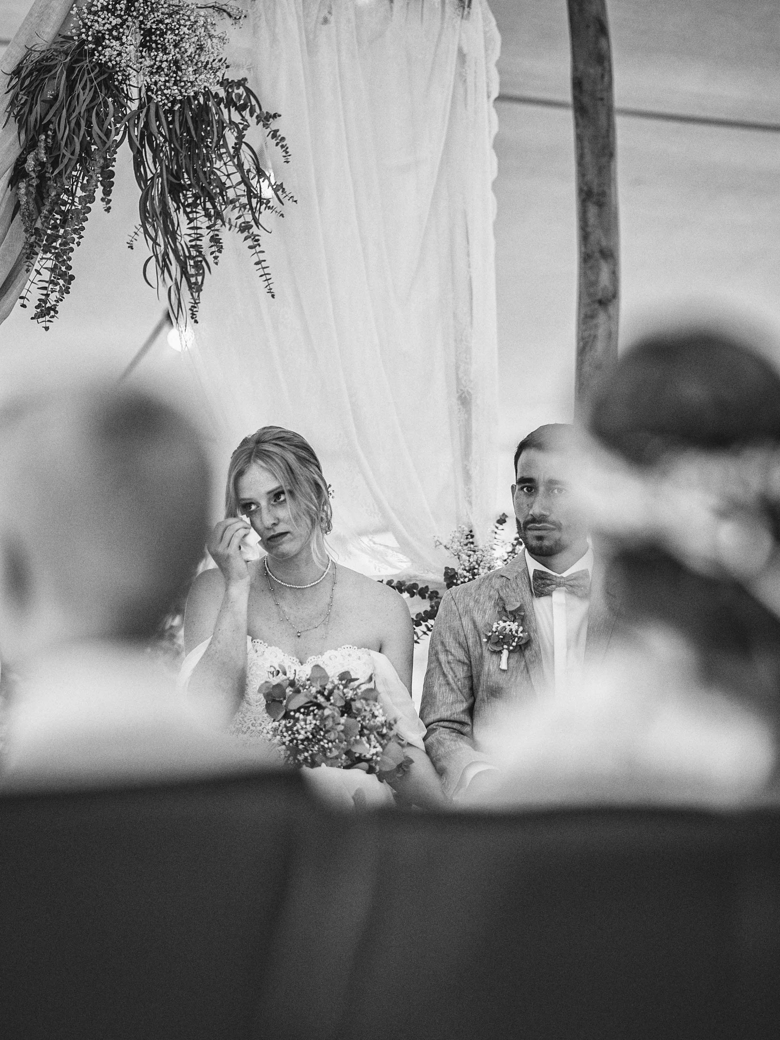 Ein schwarzig weißes Bild von einer Hochzeitszeremonie, bei der ein Brautpaar sitzt, die Braut ist emotional berührt, während der Bräutigam ernst schaut, im Vordergrund sind unscharfe Personen. Dekorative Blumen und Stoffe sind im Hintergrund sichtba