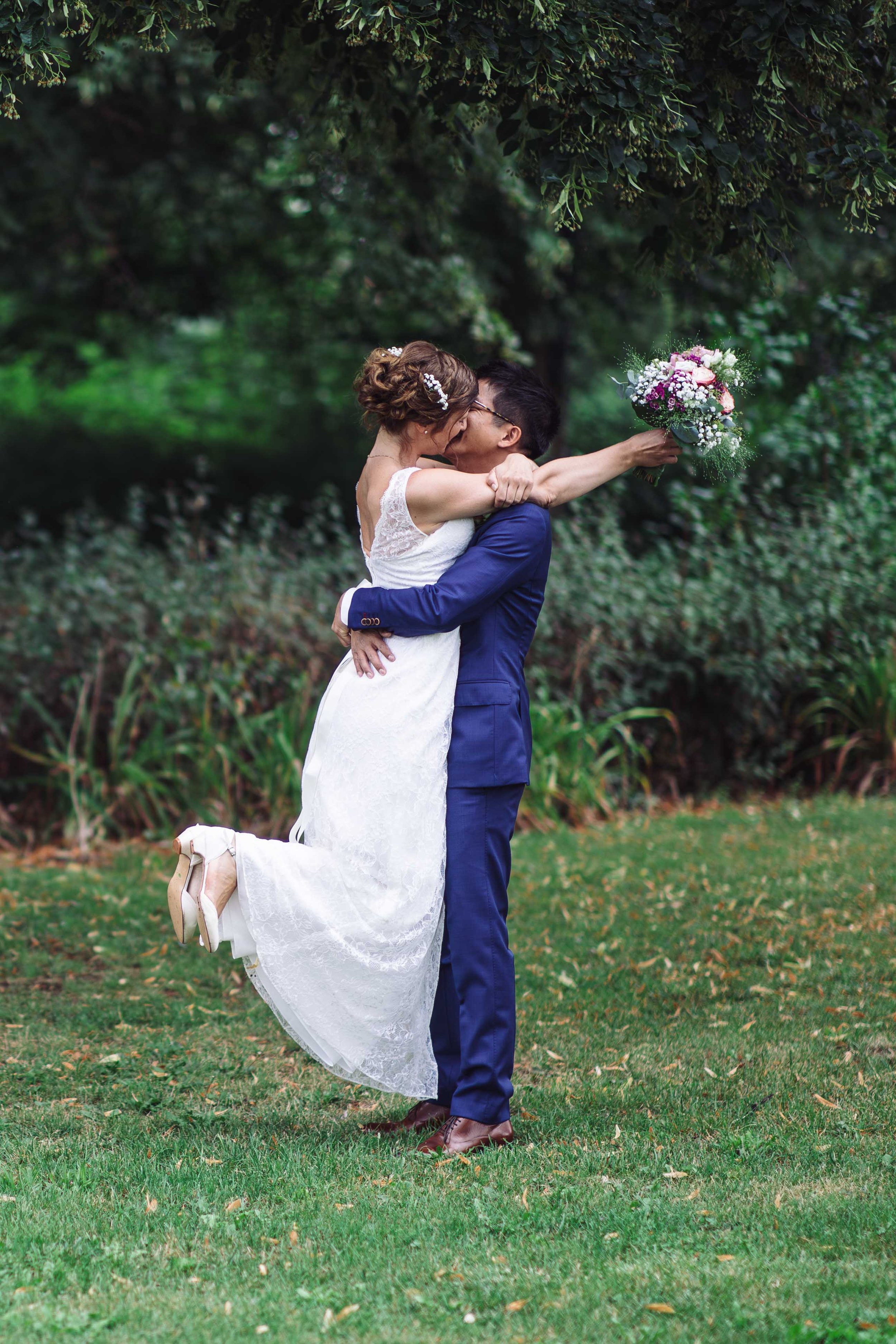 Ein Brautpaar umarmt sich und küsst sich im Park. Die Braut trägt ein weißes Hochzeitskleid und hohe Schuhe, der Bräutigam trägt einen blauen Anzug. Die Braut hält einen Blumenstrauß in der Hand.