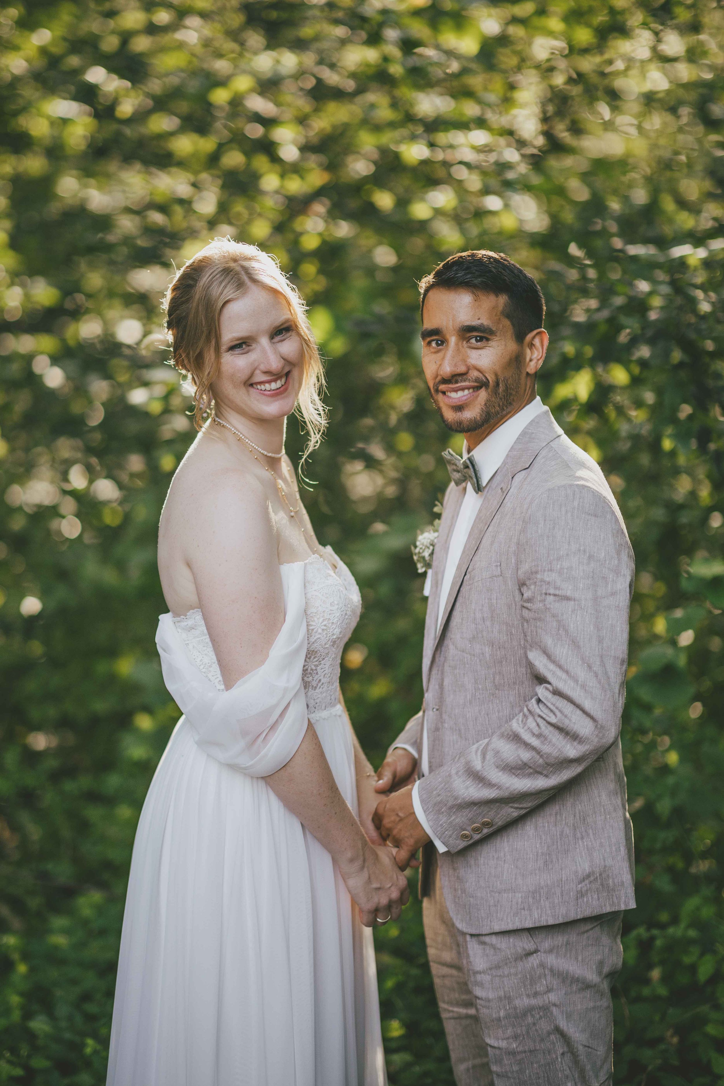 Ein lesbisches Hochzeitspaar hält Händchen in einem grünen, bewaldeten Bereich. Die Frau trägt ein weißes Hochzeitskleid, die andere einen grauen Anzug. Beide lächeln glücklich.