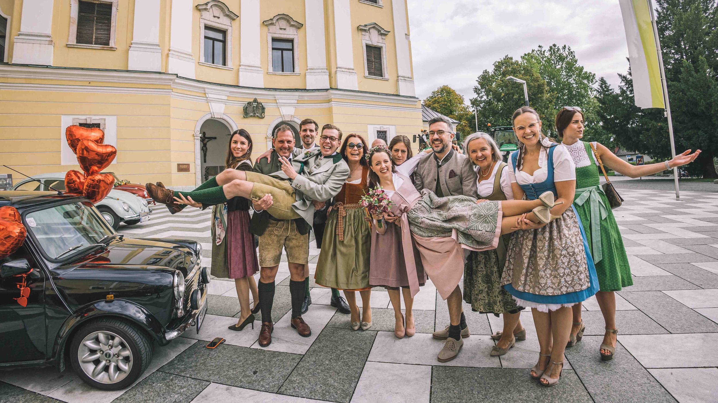 Gruppen von Menschen, Hochzeitsgästen, in traditionellen Trachten feiern vor einem gelben Gebäude, einige lachen und ltragen Braut und Bräutigam auf den Händen.