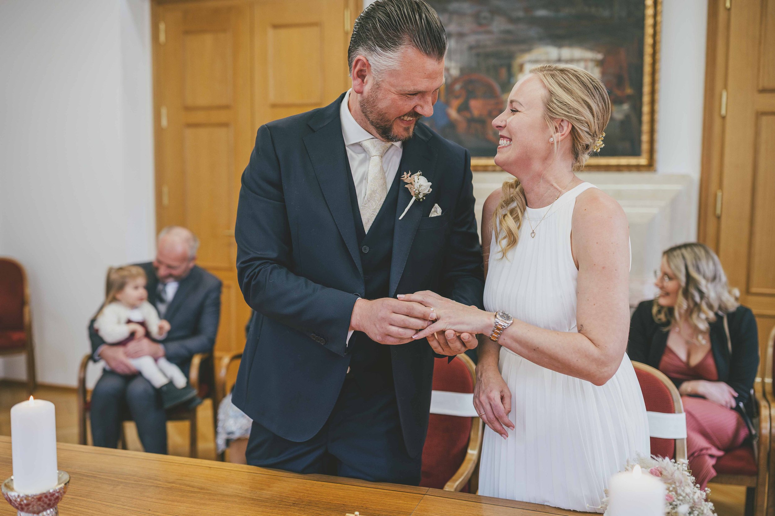 Ein Paar heiratet in einem festlichen Raum, der Bräutigam und die Braut lachen sich an und halten sich an den Händen, während sie vor Freunden und Familie stehen. Im Hintergrund sitzen weitere Gäste, darunter eine Frau mit dunklem Haar und eine junge
