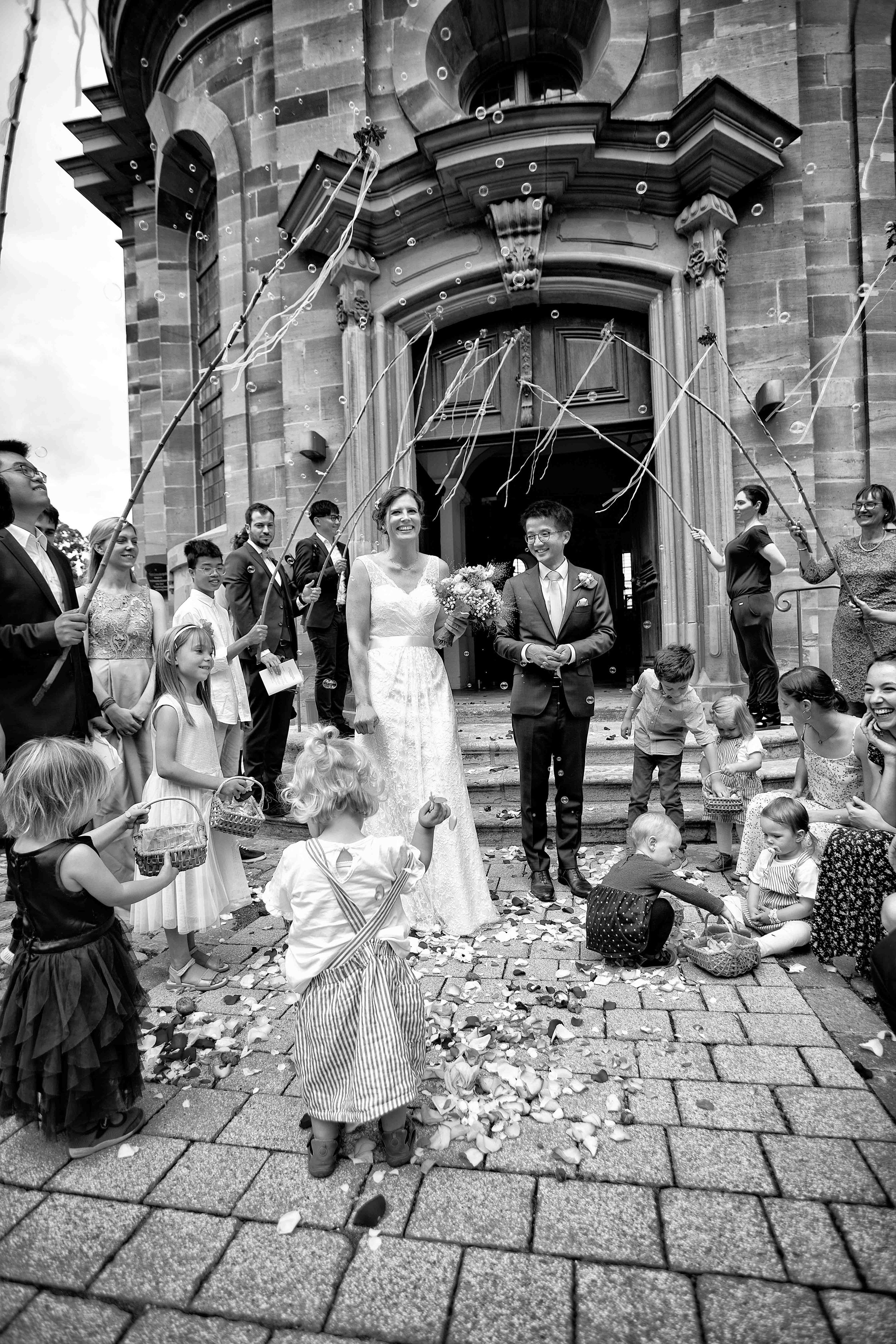 Eine schwarze und weiße Aufnahme einer Hochzeitsfeier vor einer Kirche mit vielen Kindern und Erwachsenen, die Blätter und Konfetti werfen, um das Brautpaar zu begrüßen.