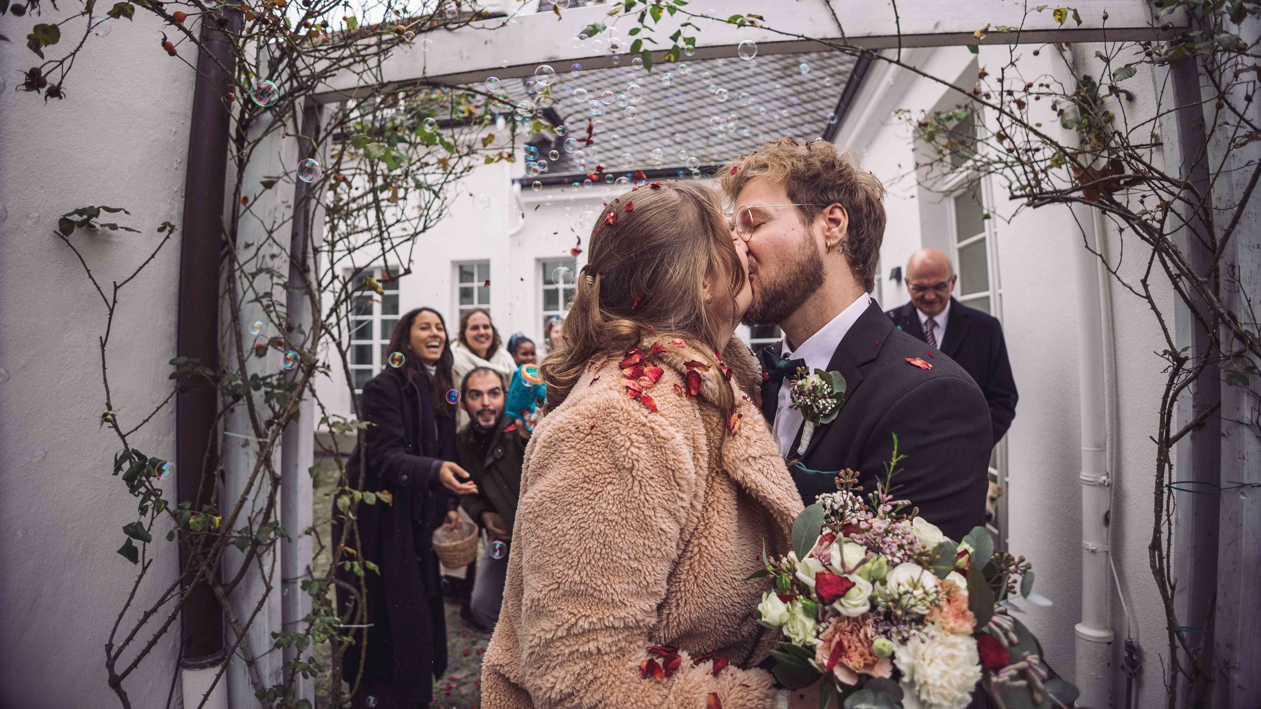 Paar bei ihrer Hochzeit küsst sich, während Freunde im Hintergrund jubeln, auf einem Weg mit blühendem Torbogen.