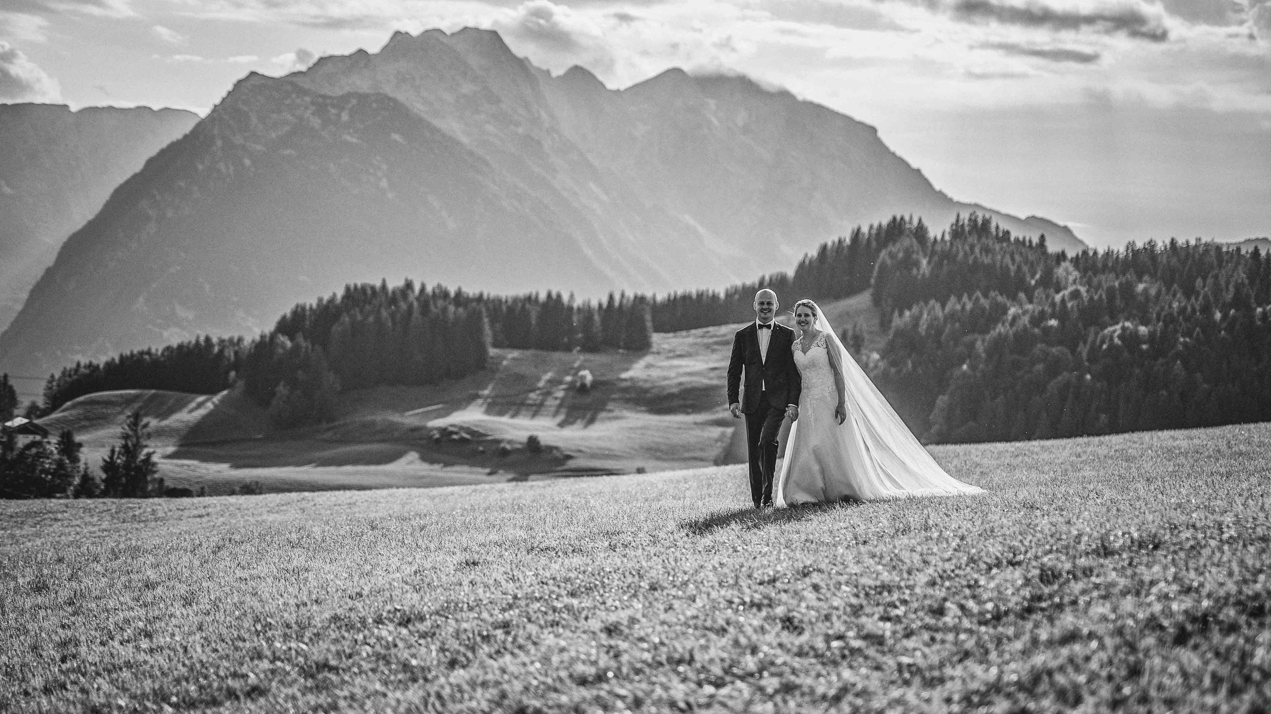 Schwarz-weiß Hochzeitsfoto eines Paares in formeller Kleidung, das auf einer Wiese vor einer Bergkulisse läuft.
