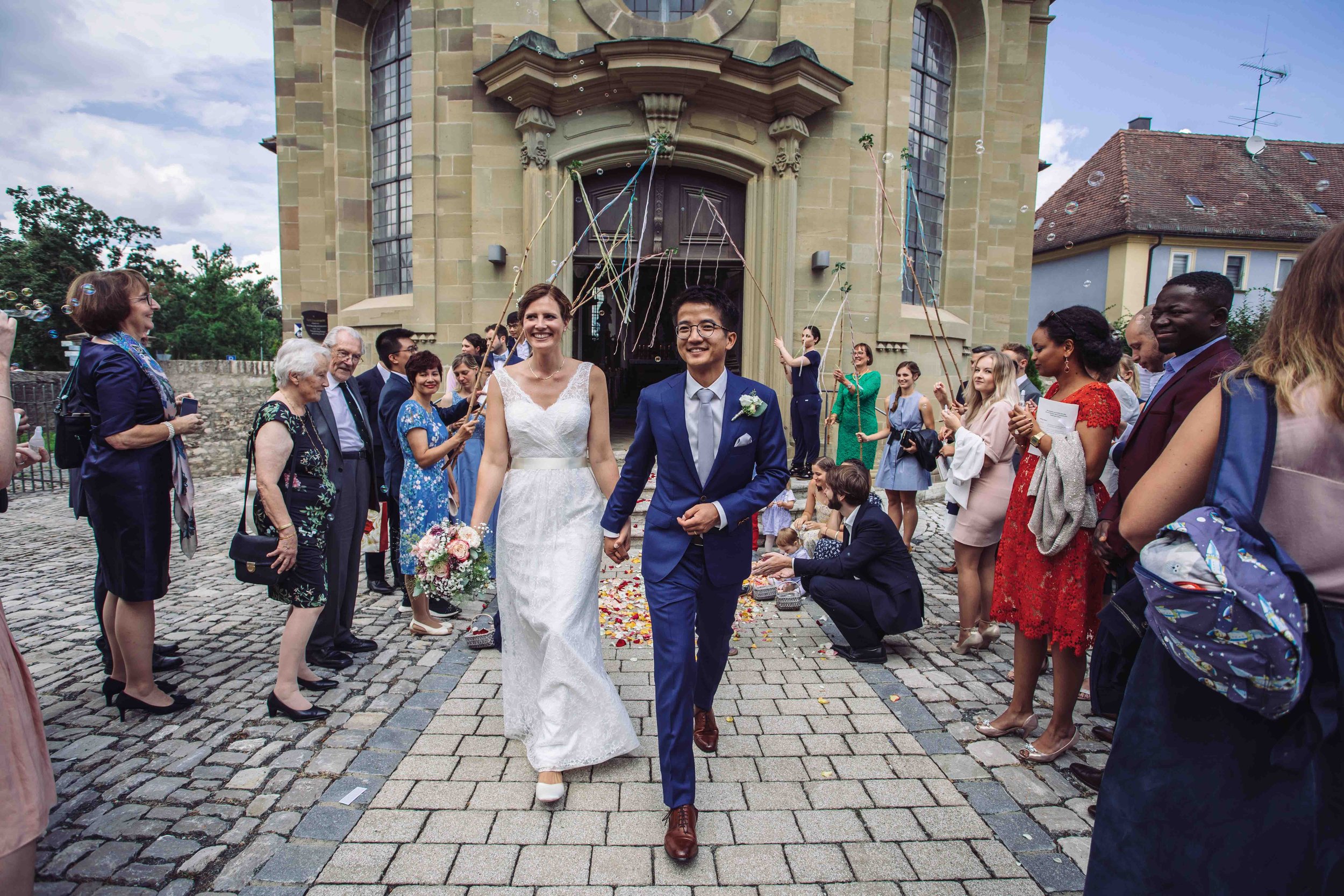 Ein glückliches Brautpaar läuft Hand in Hand aus einer Kirche, umgeben von Freunden und Familie, die ihnen gratulieren, bei einem sonnigen Hochzeitstag.