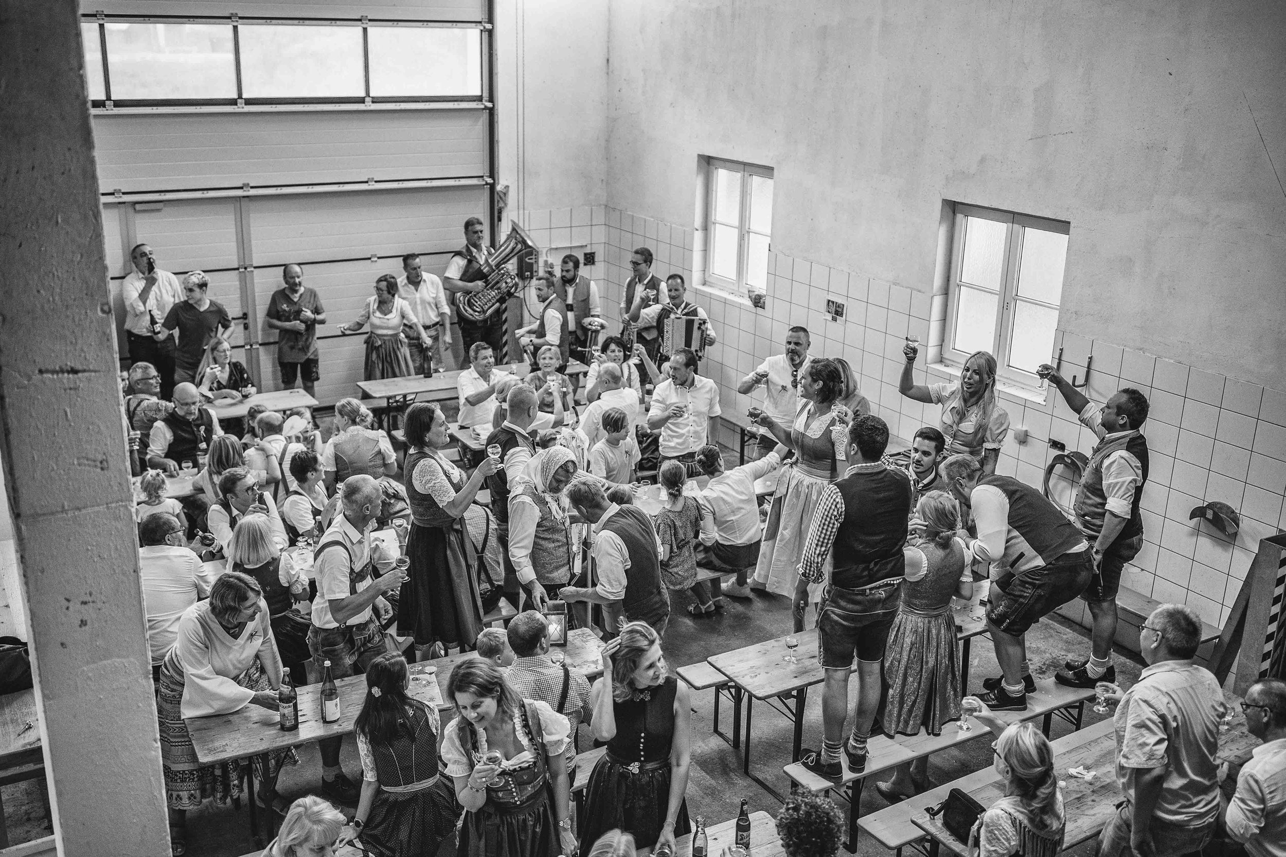 Menschen feiern in einem großen Raum, einige tanzen auf Tischen, andere sitzen an Tischen und trinken. Eine Band mit Blasinstrumenten spielt im Hintergrund, alle sind in traditioneller Kleidung.