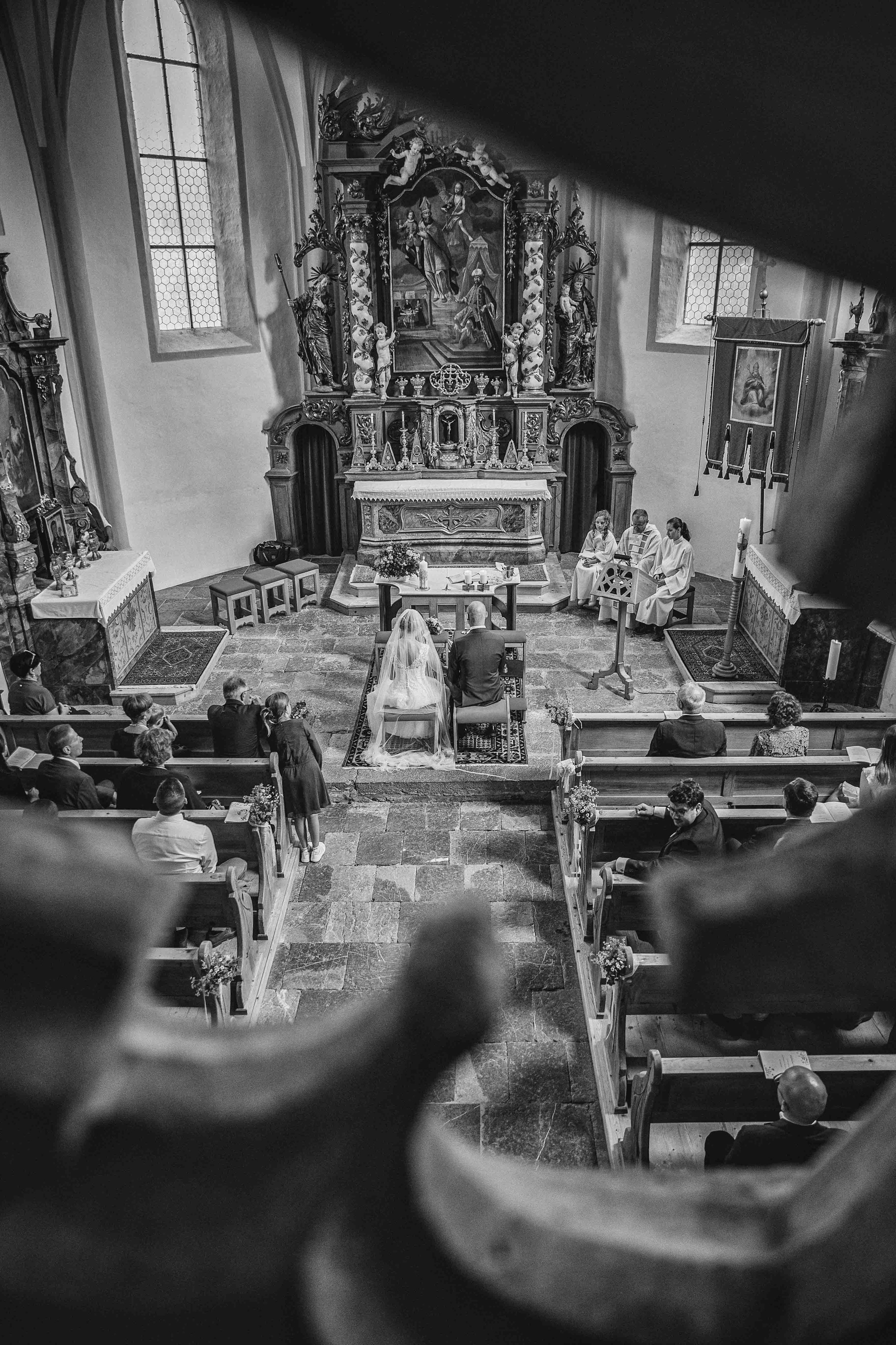 Schwarz-Weiß-Foto einer Kirchhochzeit in einer Kirche mit Hochzeitspaar, Priester, Braut in weißem Kleid und Bräutigam, Gäste sitzen auf Holzbänken, im Hintergrund ein Altar mit religiösen Statuen und Gemälden.